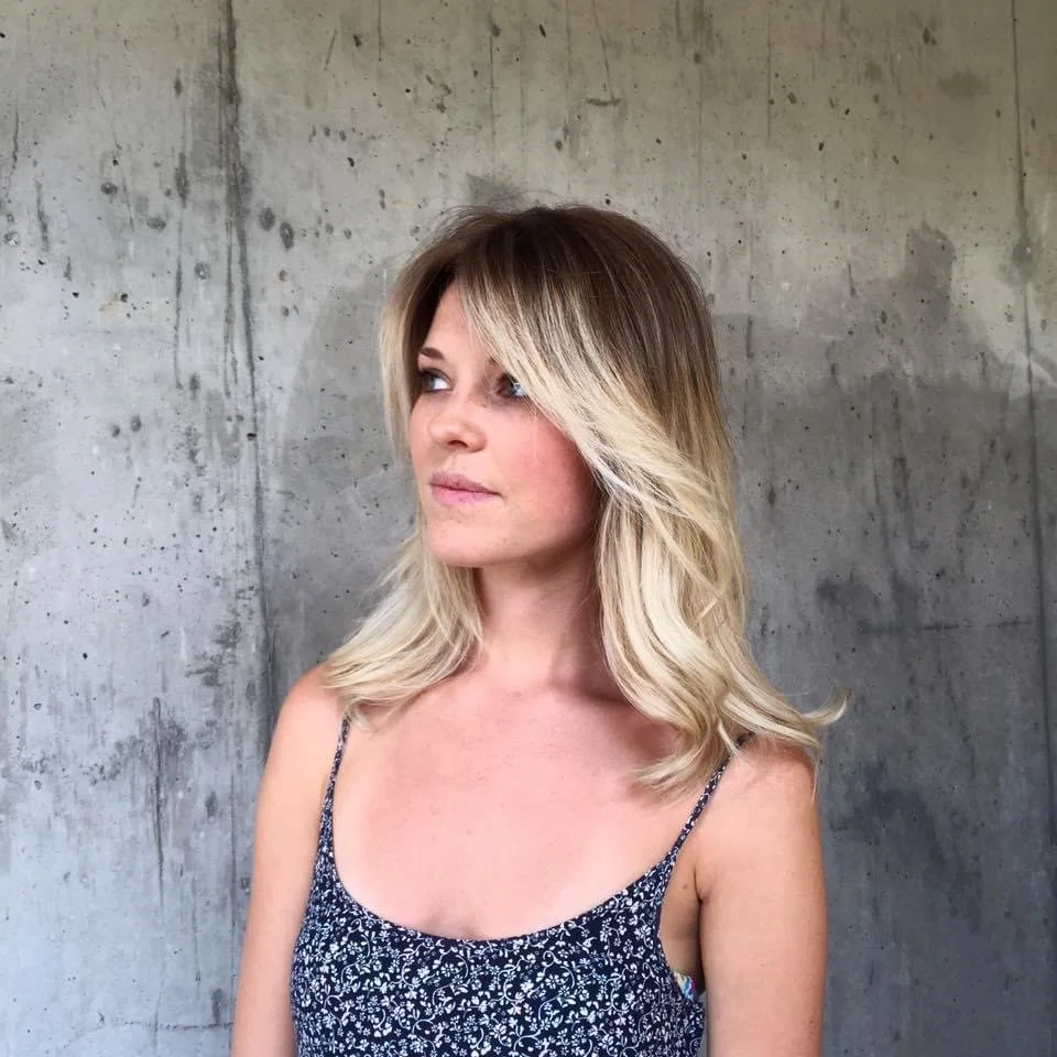 Woman with blonde layered hair looking to her left against a concrete wall background.