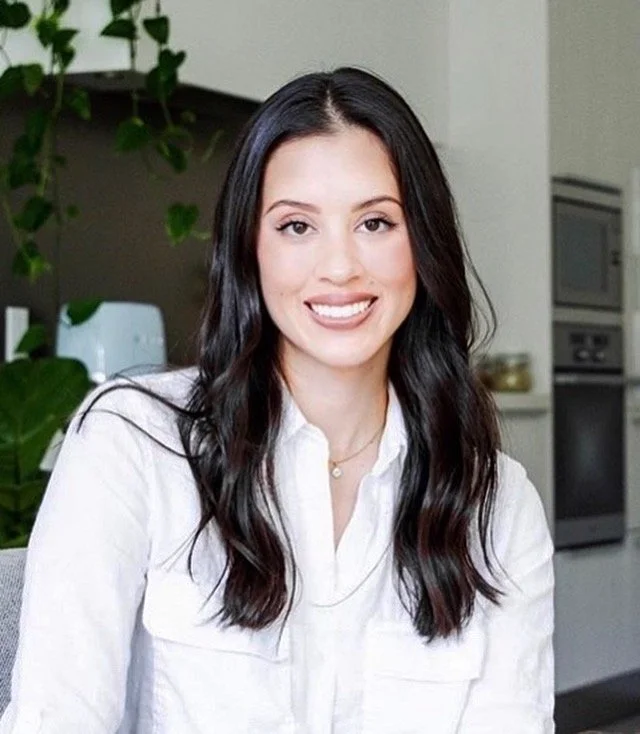 Portrait of a young woman with long dark hair, smiling, wearing a white shirt in a modern kitchen setting.