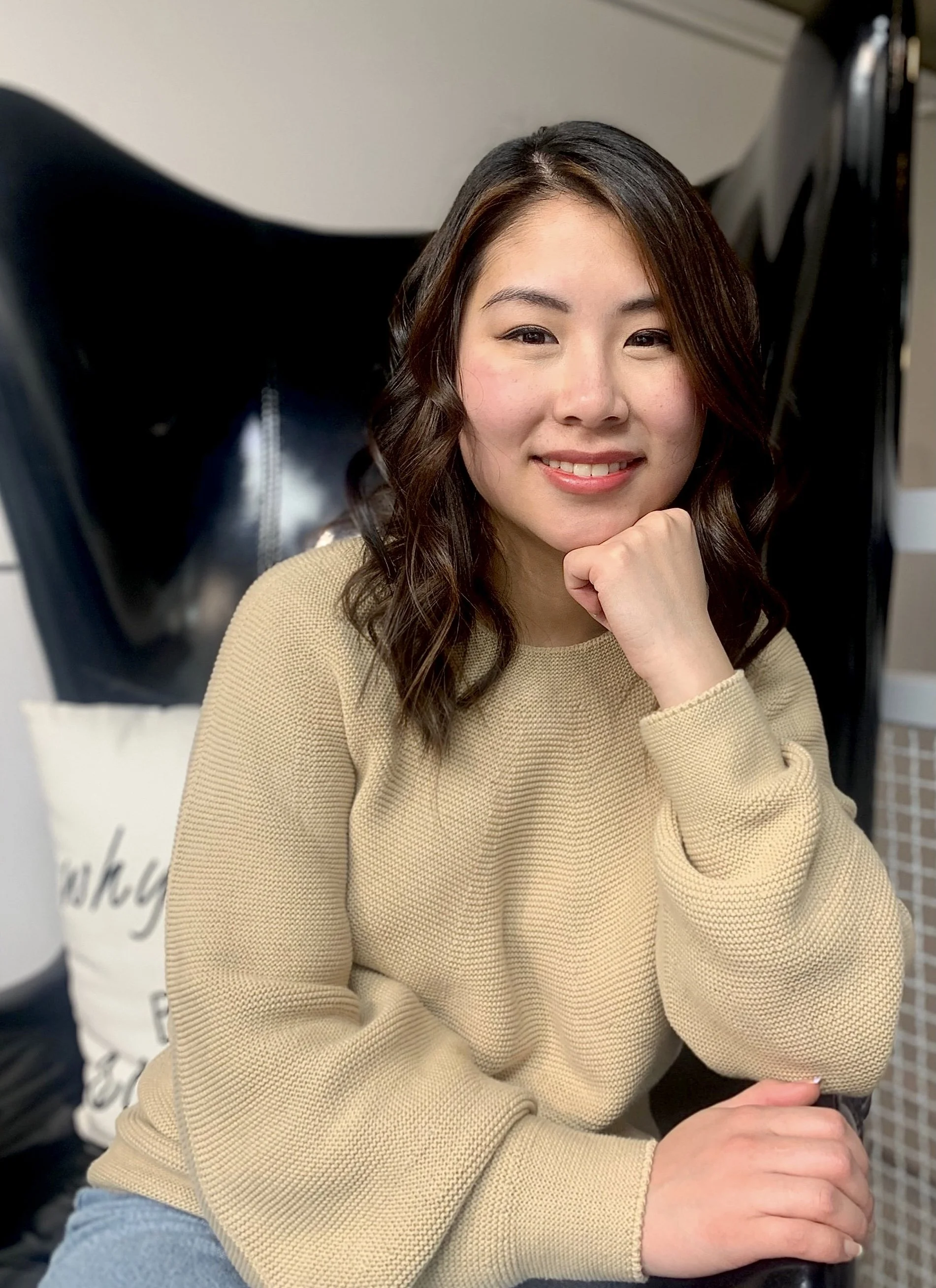 A woman in a beige sweater sitting on a chair, smiling with her hand resting on her chin. She has wavy brown hair.