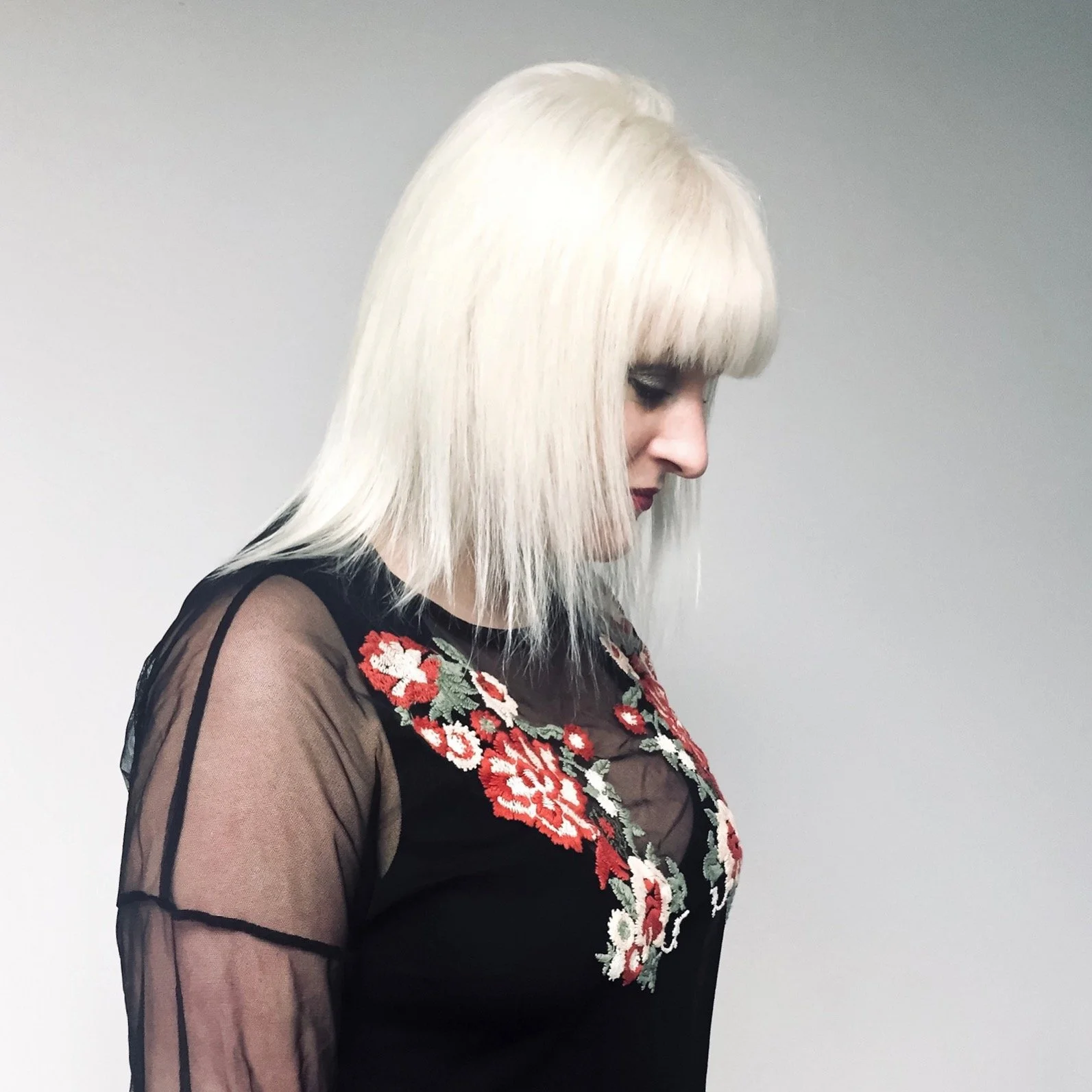 Side profile of a woman with platinum blonde hair in a bob cut, wearing a black sheer top with red and white floral embroidery.