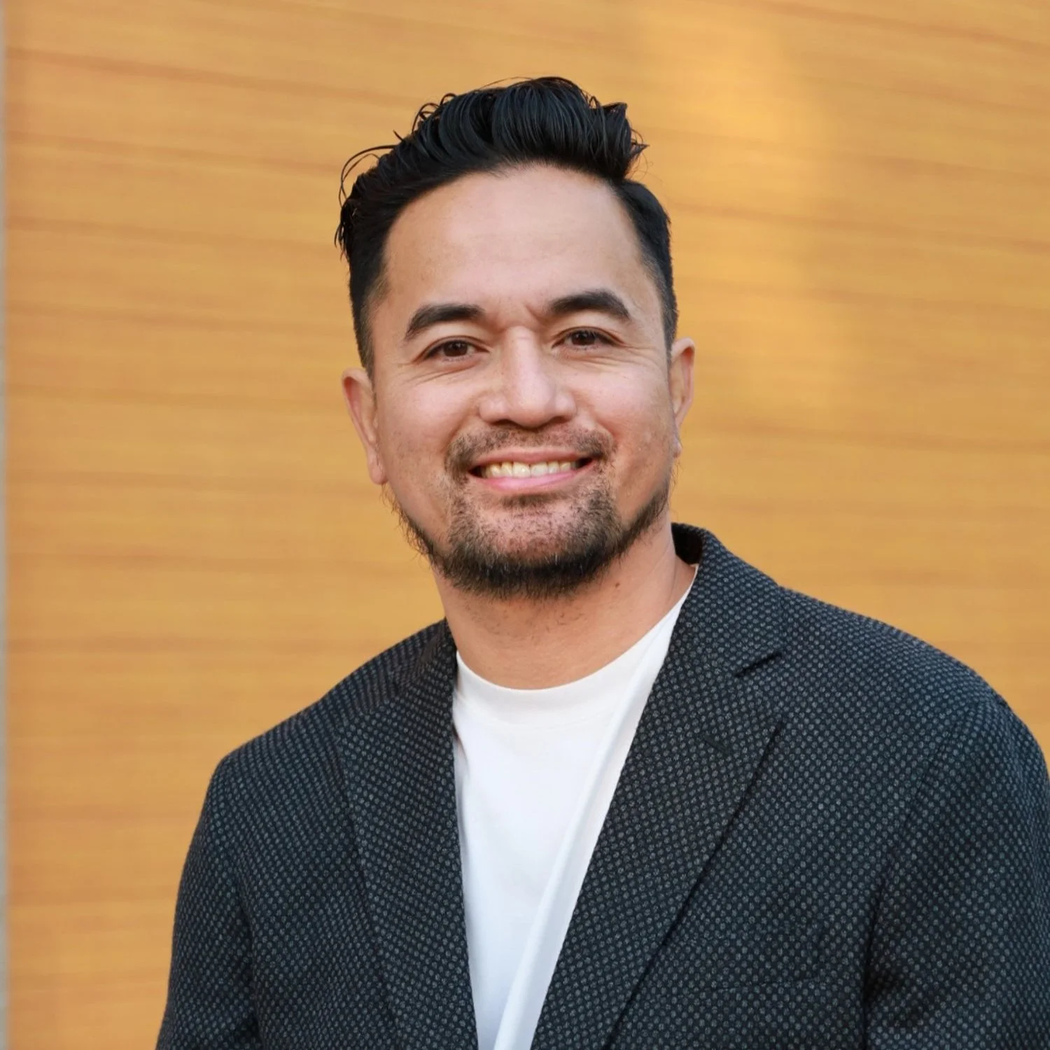 Man in a dark suit jacket smiling in front of a wooden background.