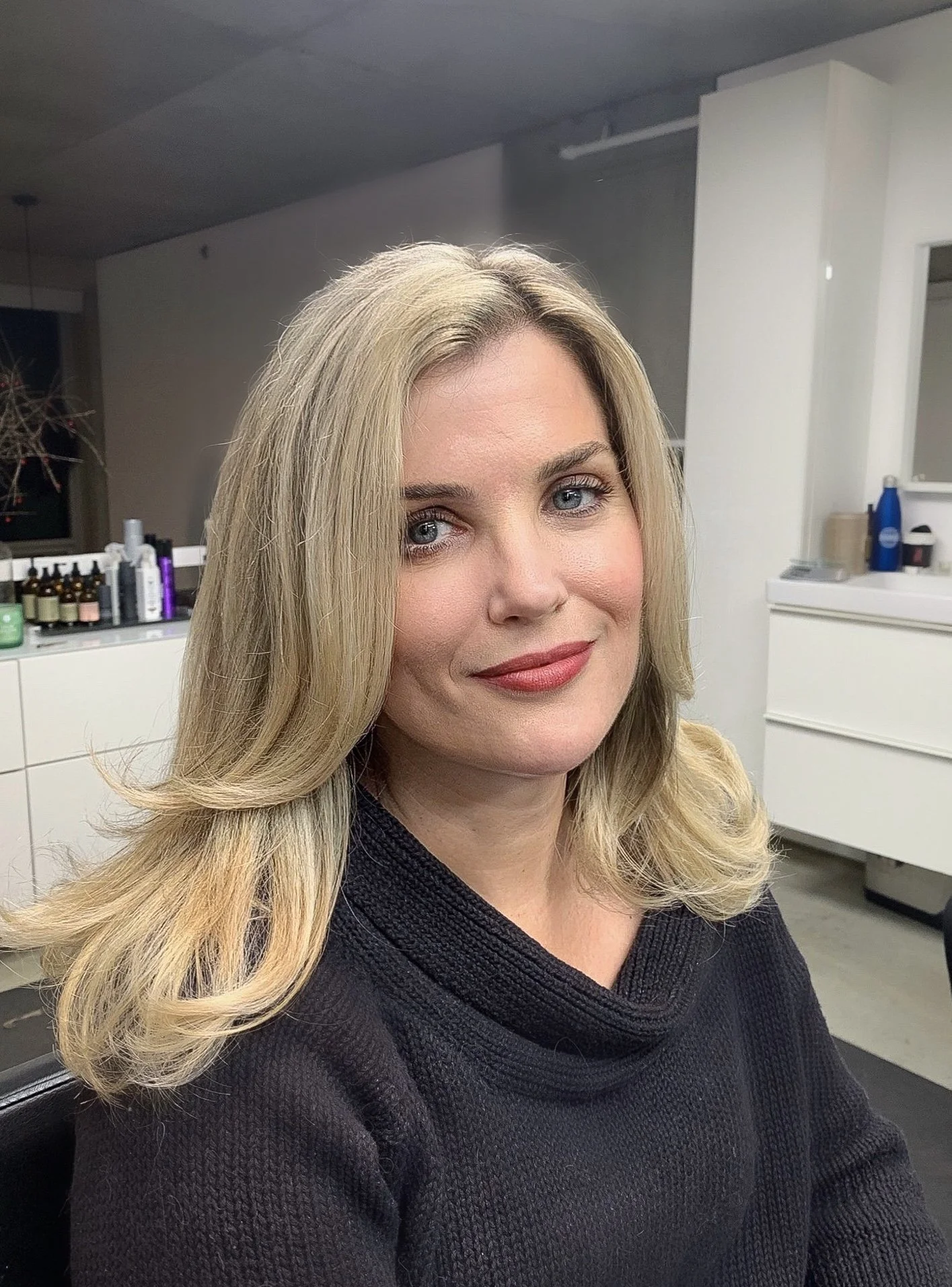 A woman with blonde hair and blue eyes smiling, seated in a salon or similar setting.