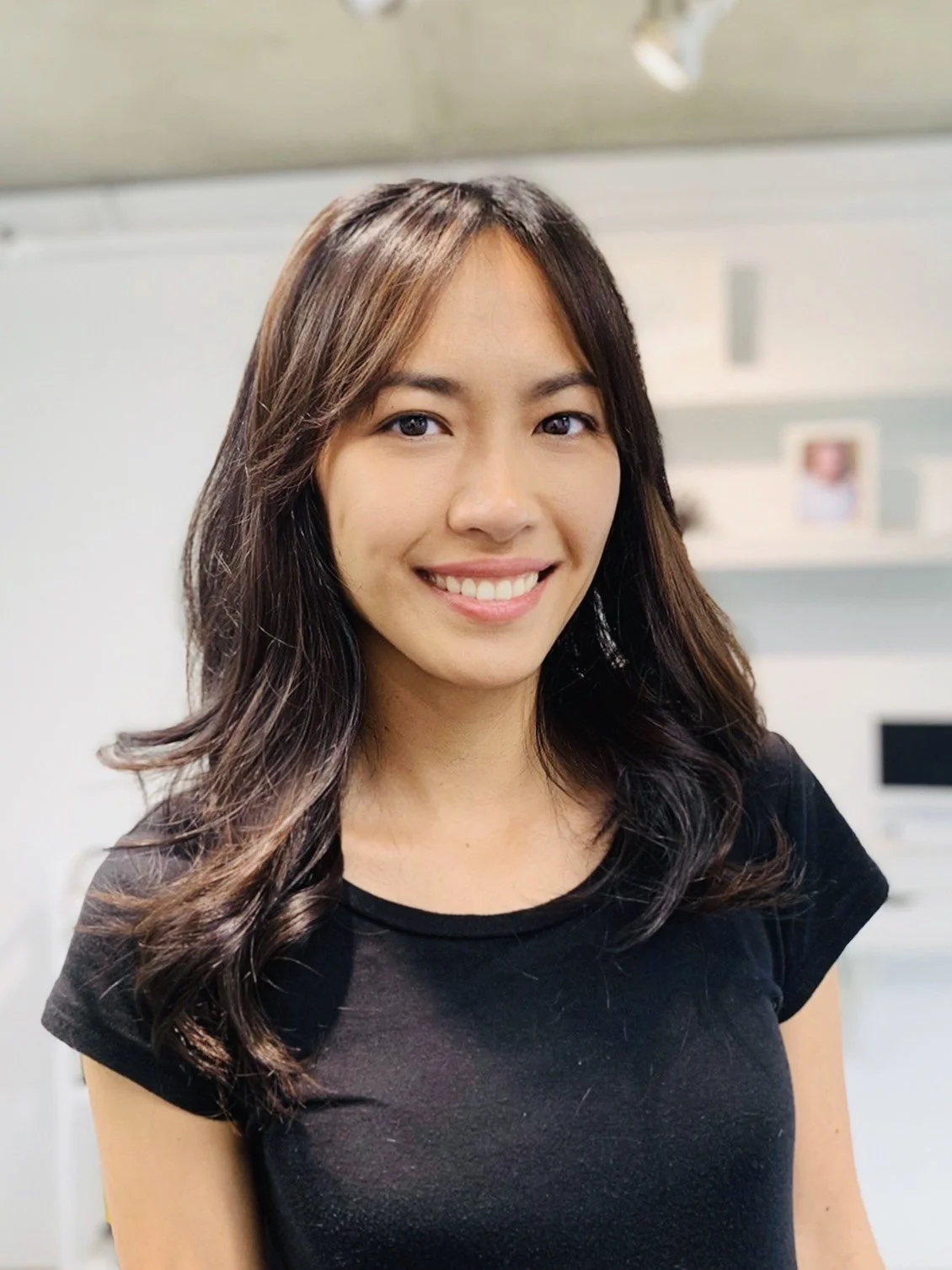 Smiling person with long dark hair wearing a black shirt, standing indoors.