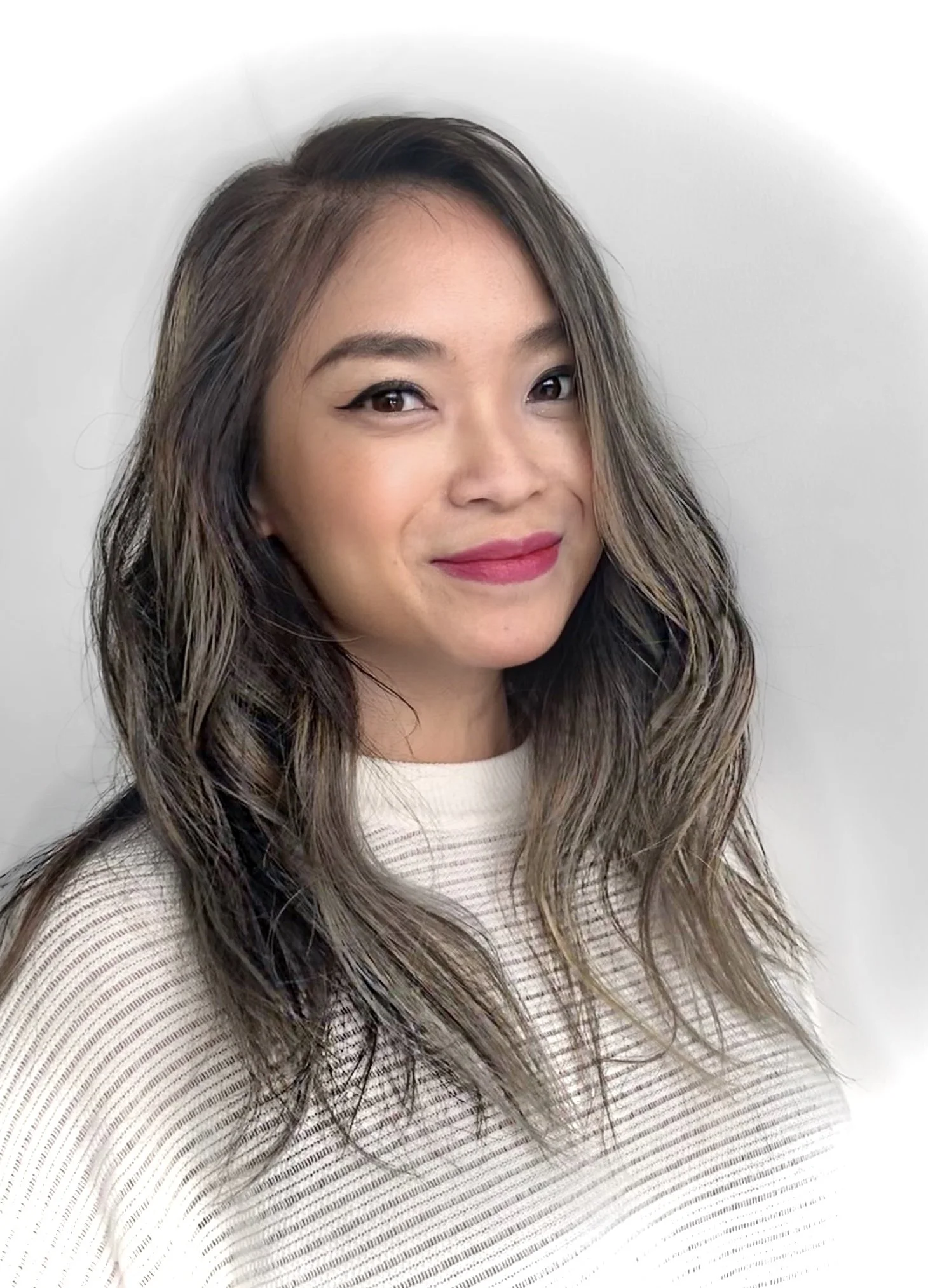 Portrait of a young woman with wavy brown hair, wearing a cream-colored top, smiling with pink lipstick, against a plain white background.
