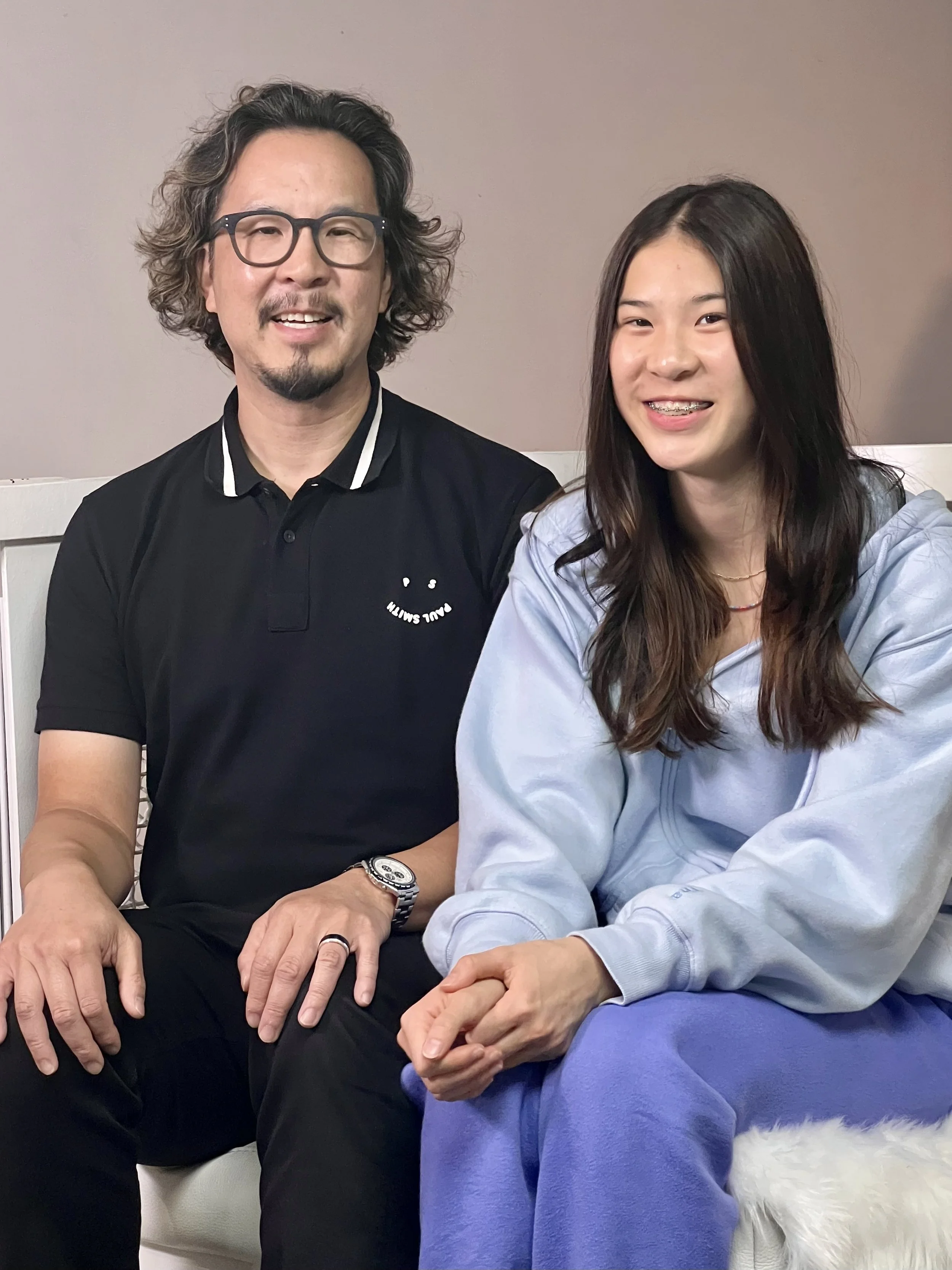 Two people, a man and a woman, sitting together indoors smiling. The man is wearing glasses and a black polo shirt, while the woman is in a light blue sweatshirt.