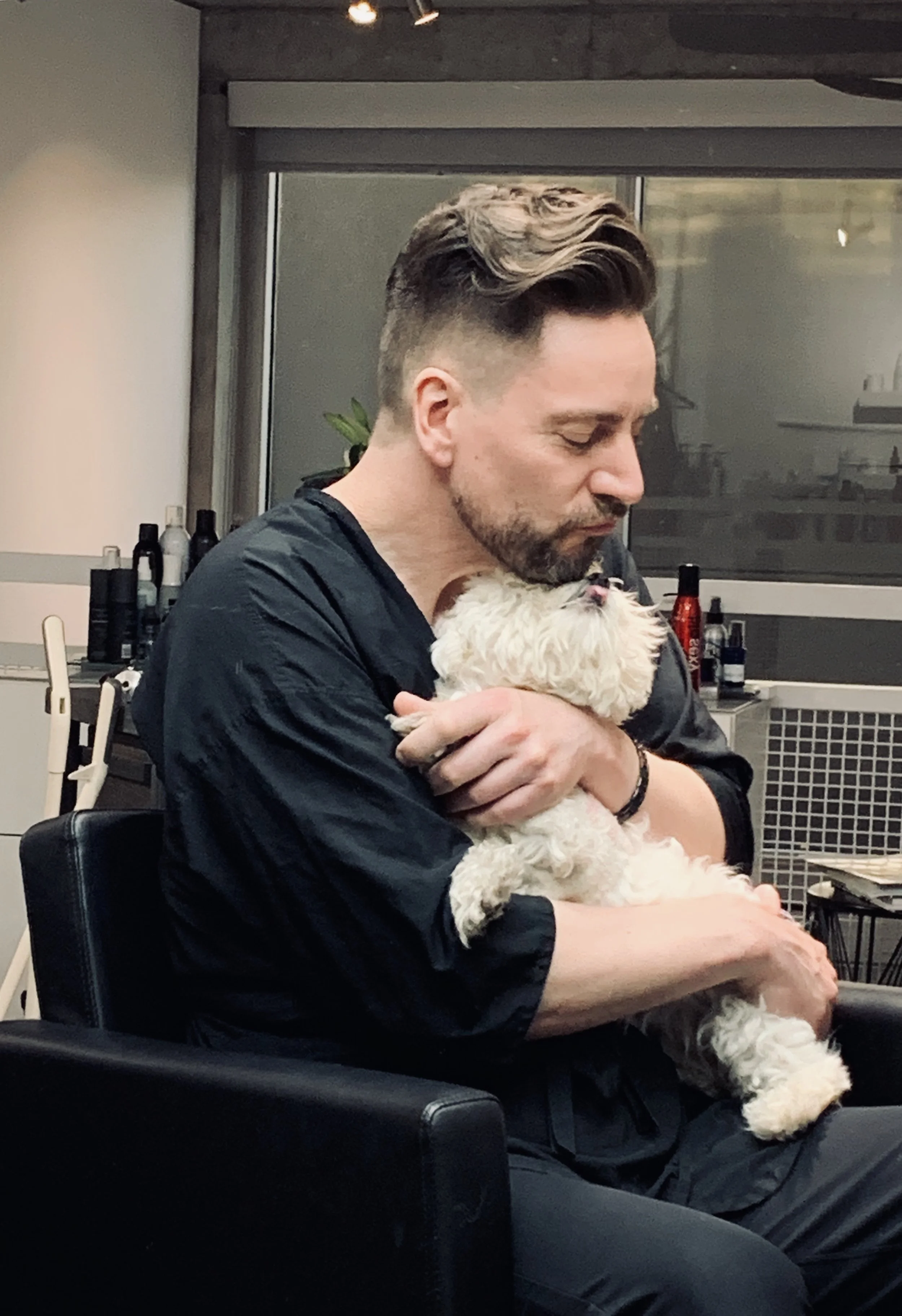 Man sitting in a chair hugging a small white dog in a room with shelves and bottles.
