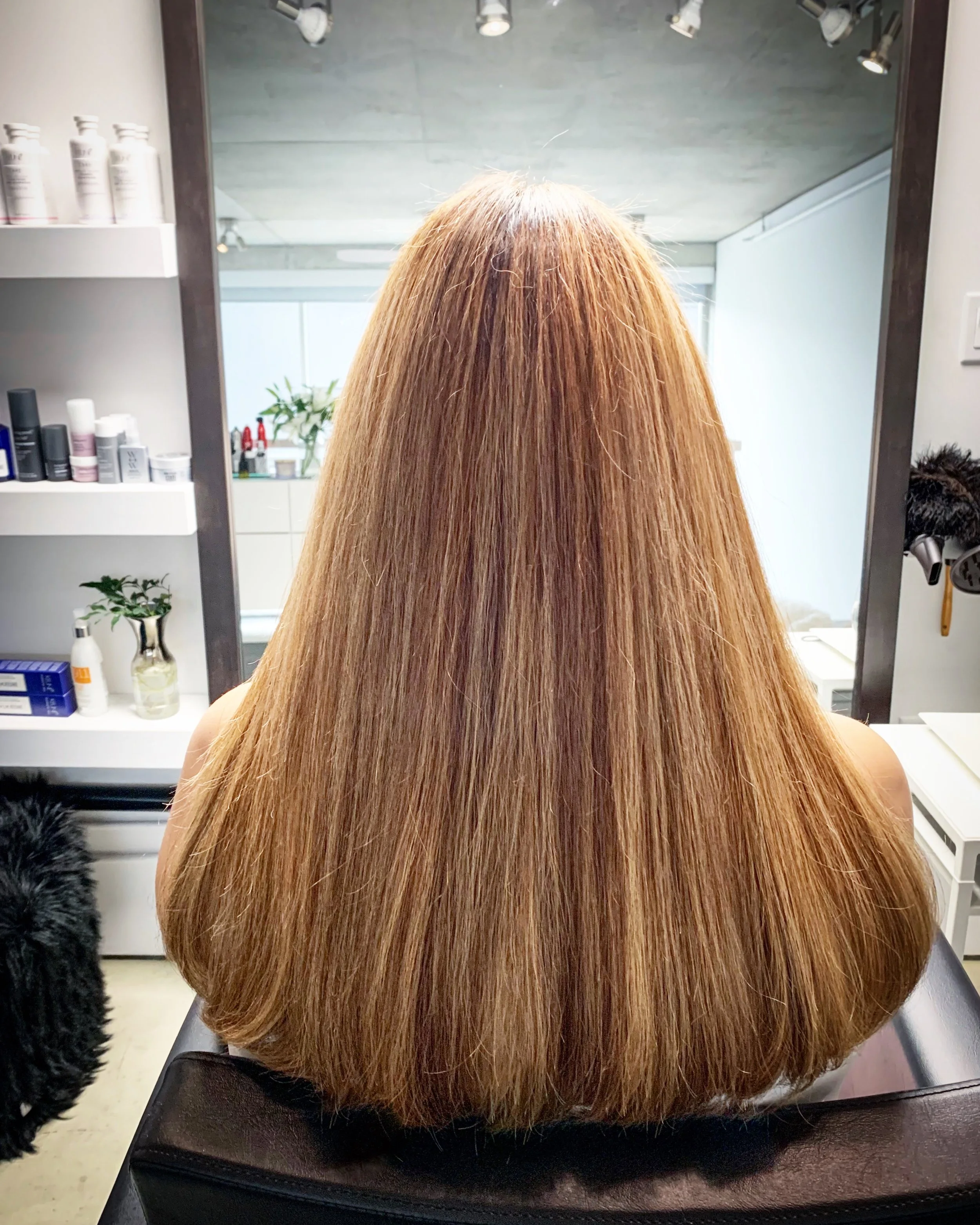 Back view of a person with straight, medium-length blonde hair in a salon setting, facing a mirror with hair products visible on the shelves.