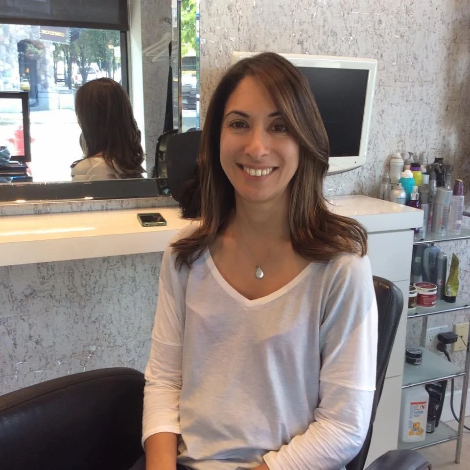 A woman with shoulder-length brown hair and a white long-sleeve shirt is sitting on a black salon chair, smiling. Behind her, there's a mirror, a wall with a textured surface, a small TV, and a shelf with hair and beauty products.