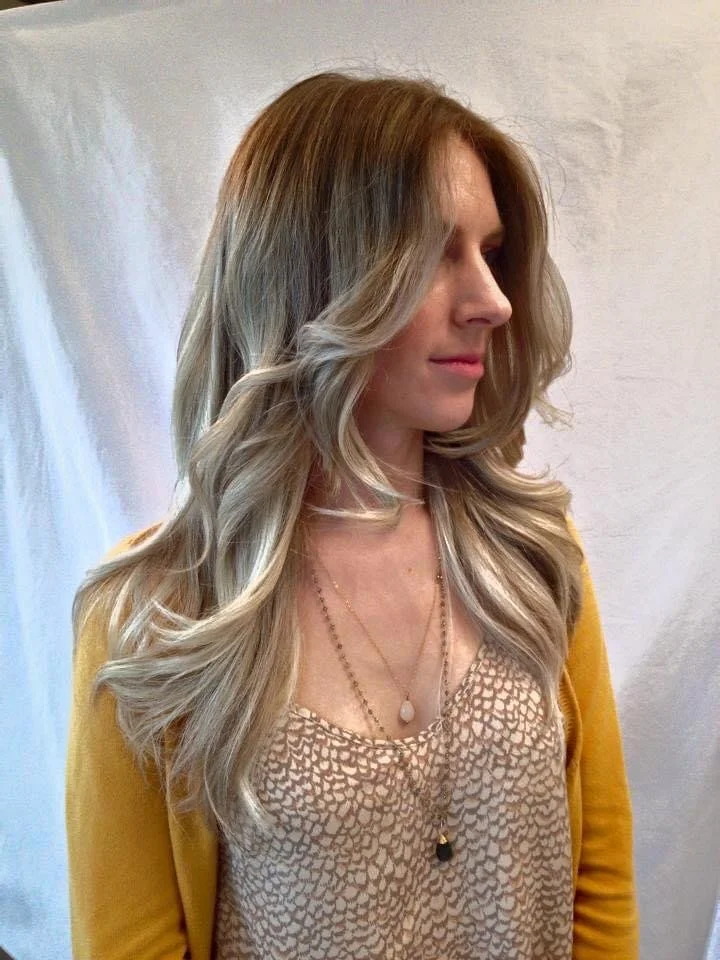 Side profile of a woman with long, wavy blonde hair, wearing a beige top with a pattern and a yellow cardigan, accessorized with layered necklaces, standing against a light-colored wall.