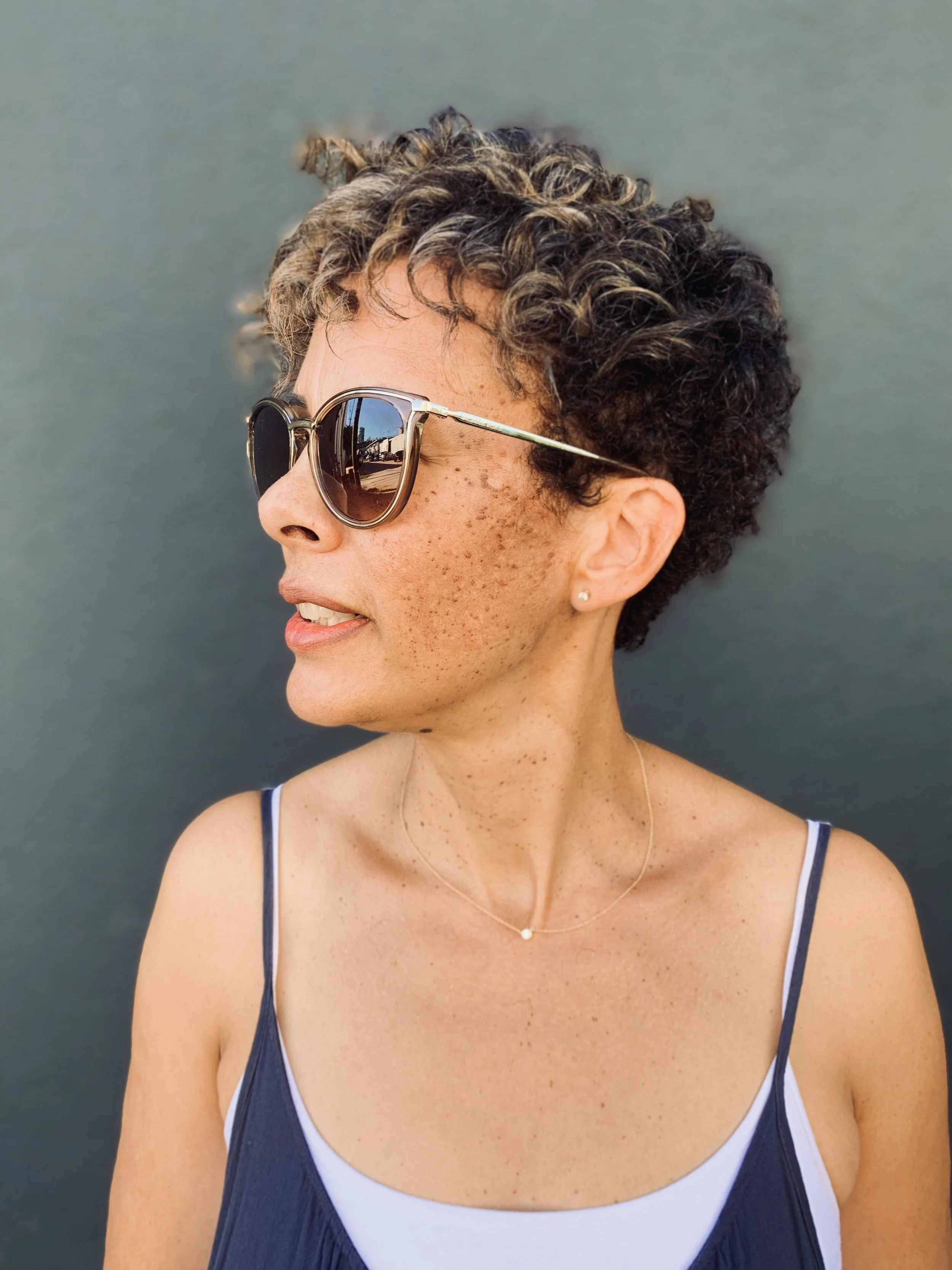 Woman with curly hair wearing sunglasses and a sleeveless top, looking to the side, against a gray background.