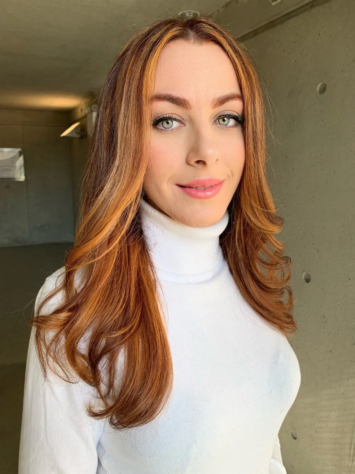 A woman with long, wavy red hair and blue eyes wearing a white turtleneck sweater, standing indoors against a concrete wall.