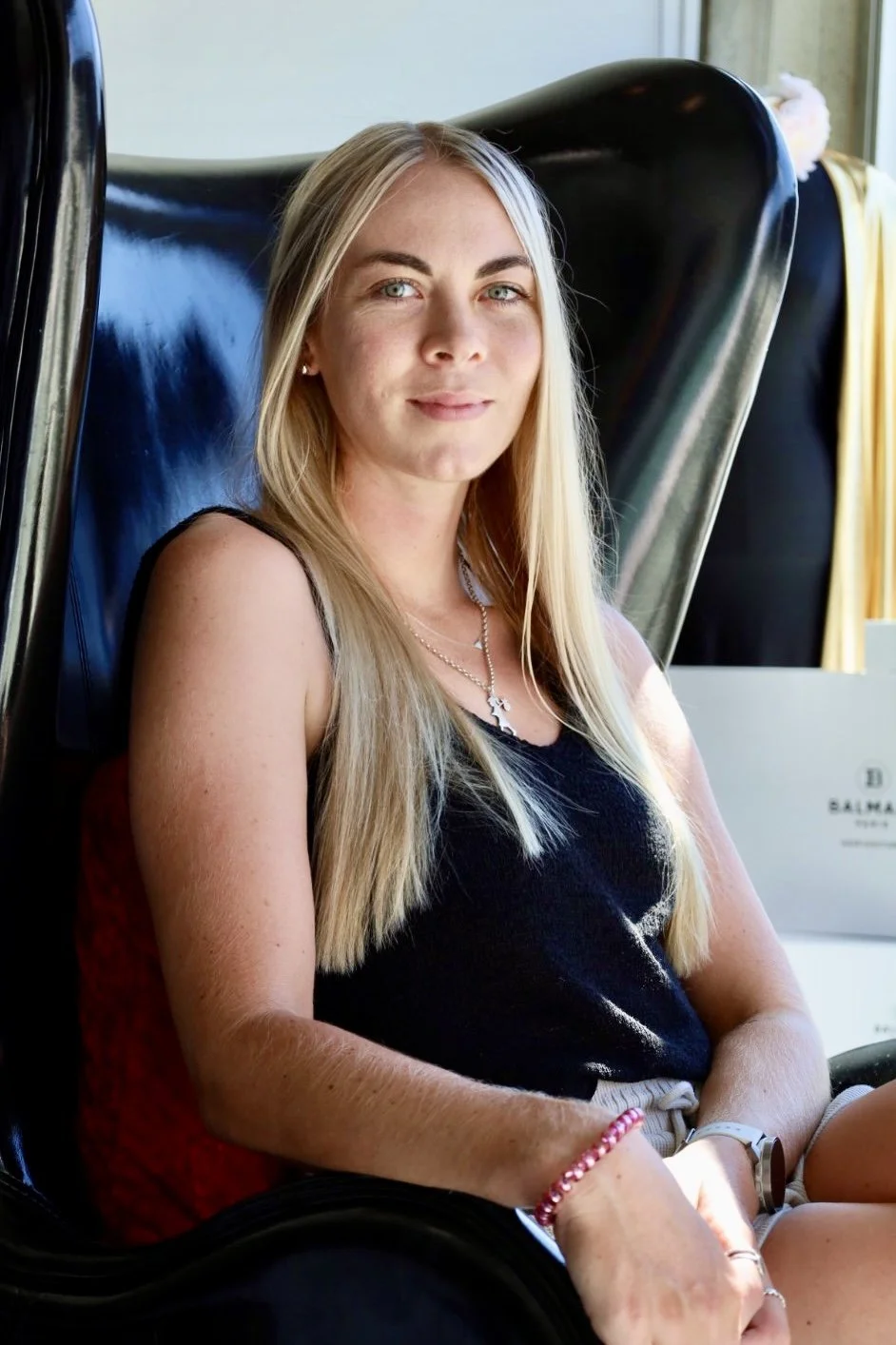 Woman sitting in a black chair, wearing a black top and necklace with a cross, with long blonde hair and neutral expression.