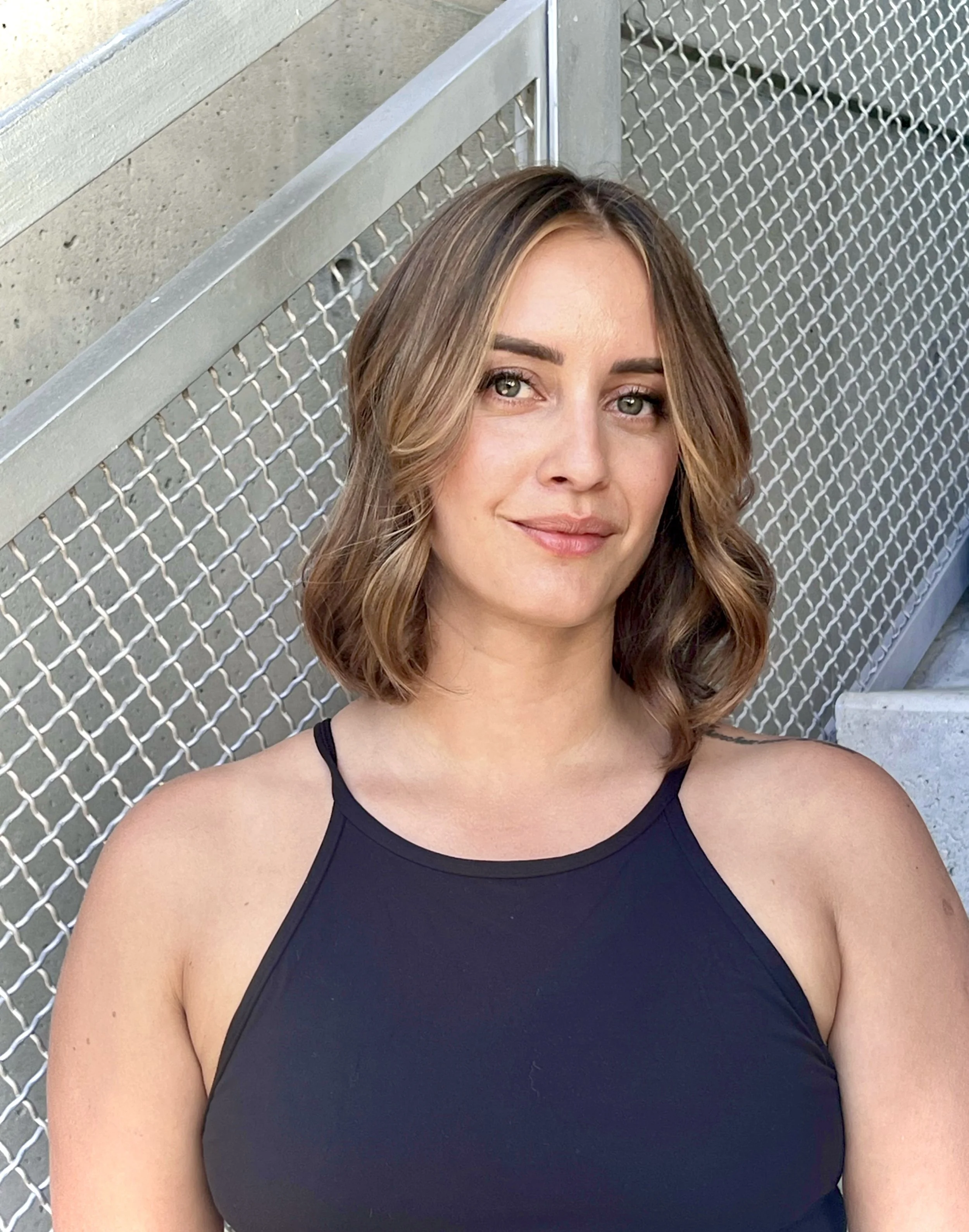 Person with shoulder-length hair wearing a black sleeveless top, standing in front of a wire fence and concrete wall.