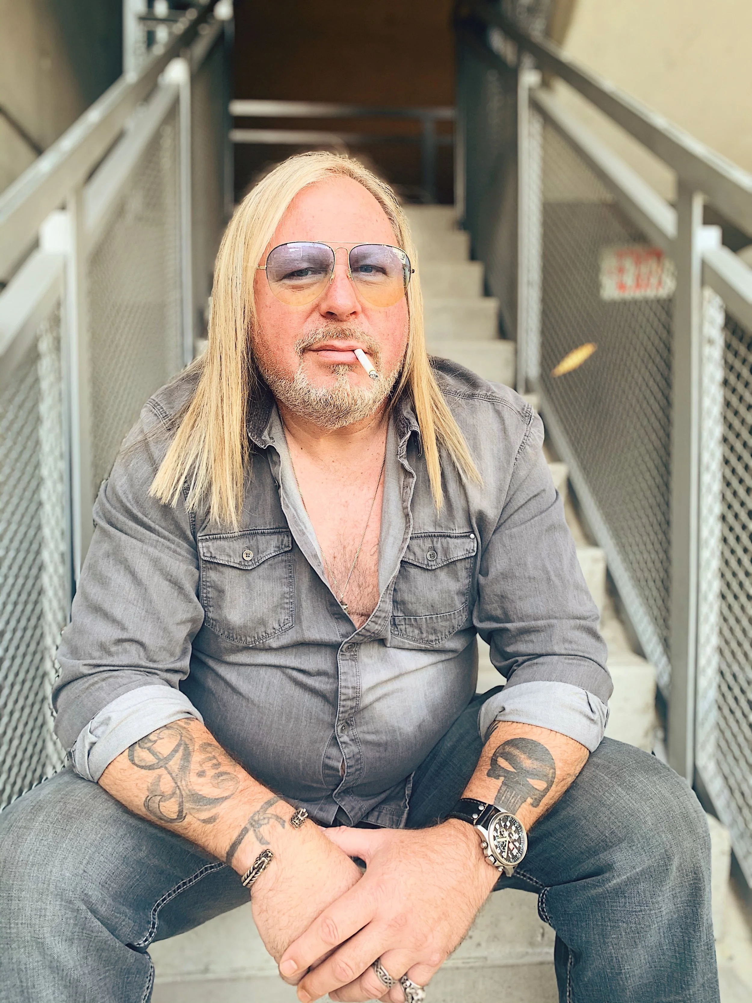A man with long blond hair, a beard, and sunglasses sitting on stairs, holding a cigarette in his mouth, wearing a gray shirt with rolled-up sleeves, tattoos on arms, and jewelry including rings, a bracelet, and a watch.
