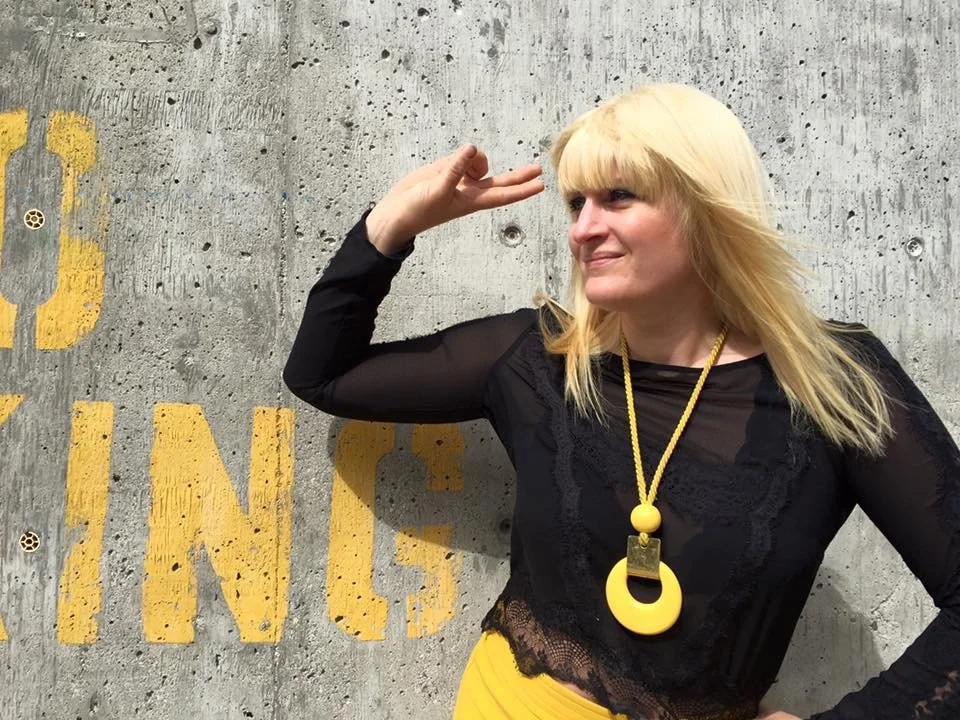 A woman with blonde hair saluting against a concrete wall with yellow painted text.