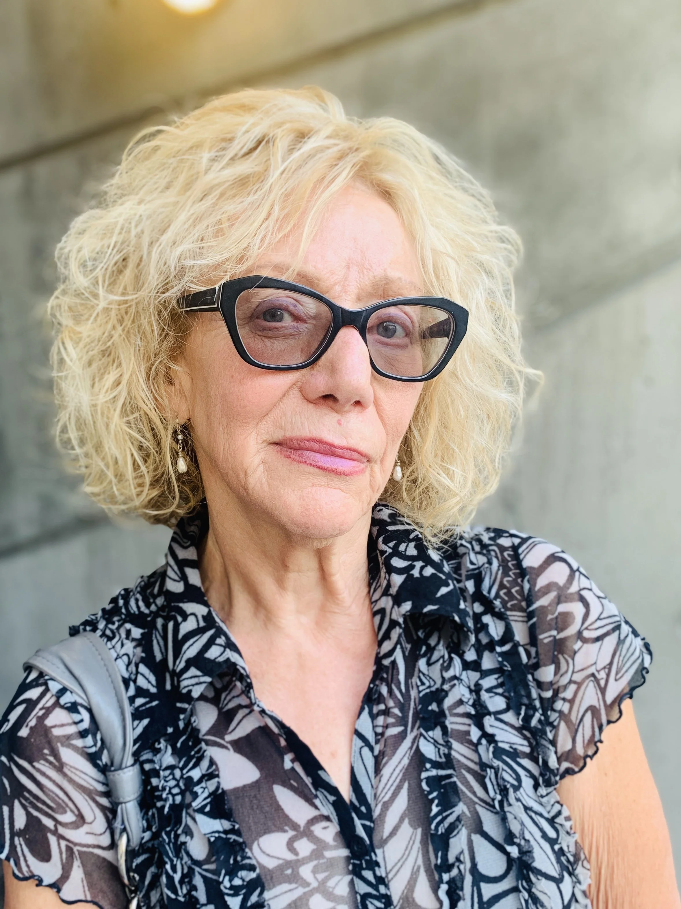 Elderly woman with curly blonde hair and black-rimmed glasses, wearing a patterned blouse.