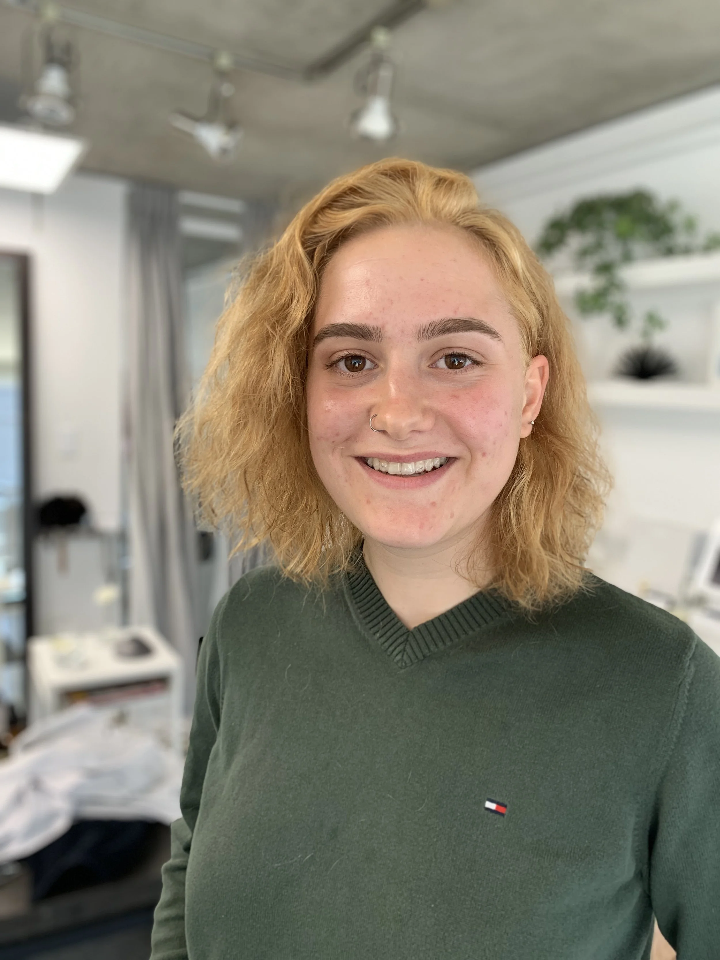 Smiling person with curly blond hair in a green sweater, standing indoors with a modern white and gray background.