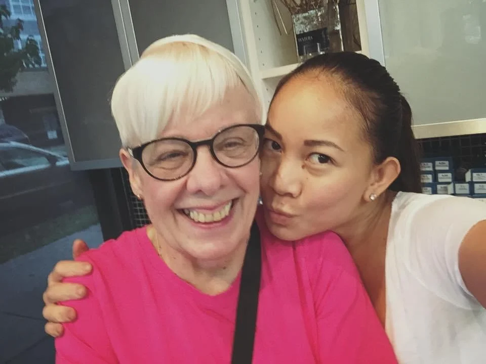 Two women taking a selfie together indoors. The woman on the left has short blonde hair, glasses, and is wearing a pink top, smiling broadly. The woman on the right has dark hair tied back, is making a kiss face, and is wearing a white top.