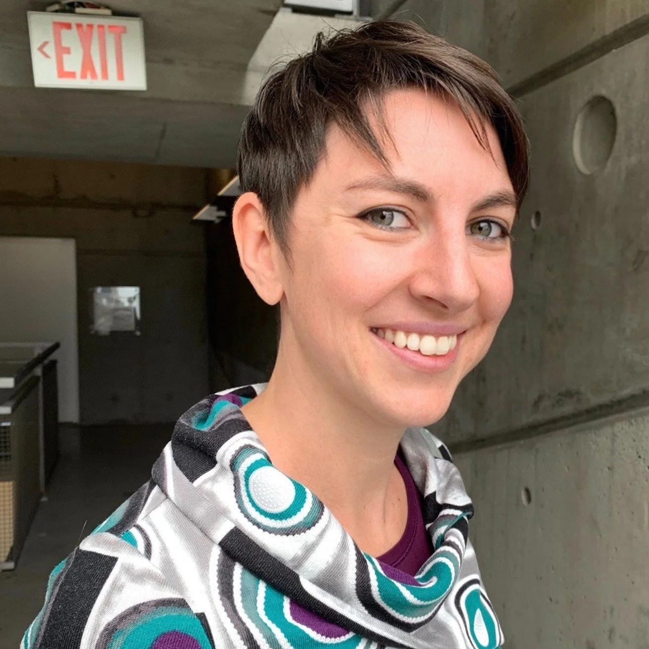 Smiling woman with short dark hair in an urban or industrial setting, wearing a patterned hoodie.