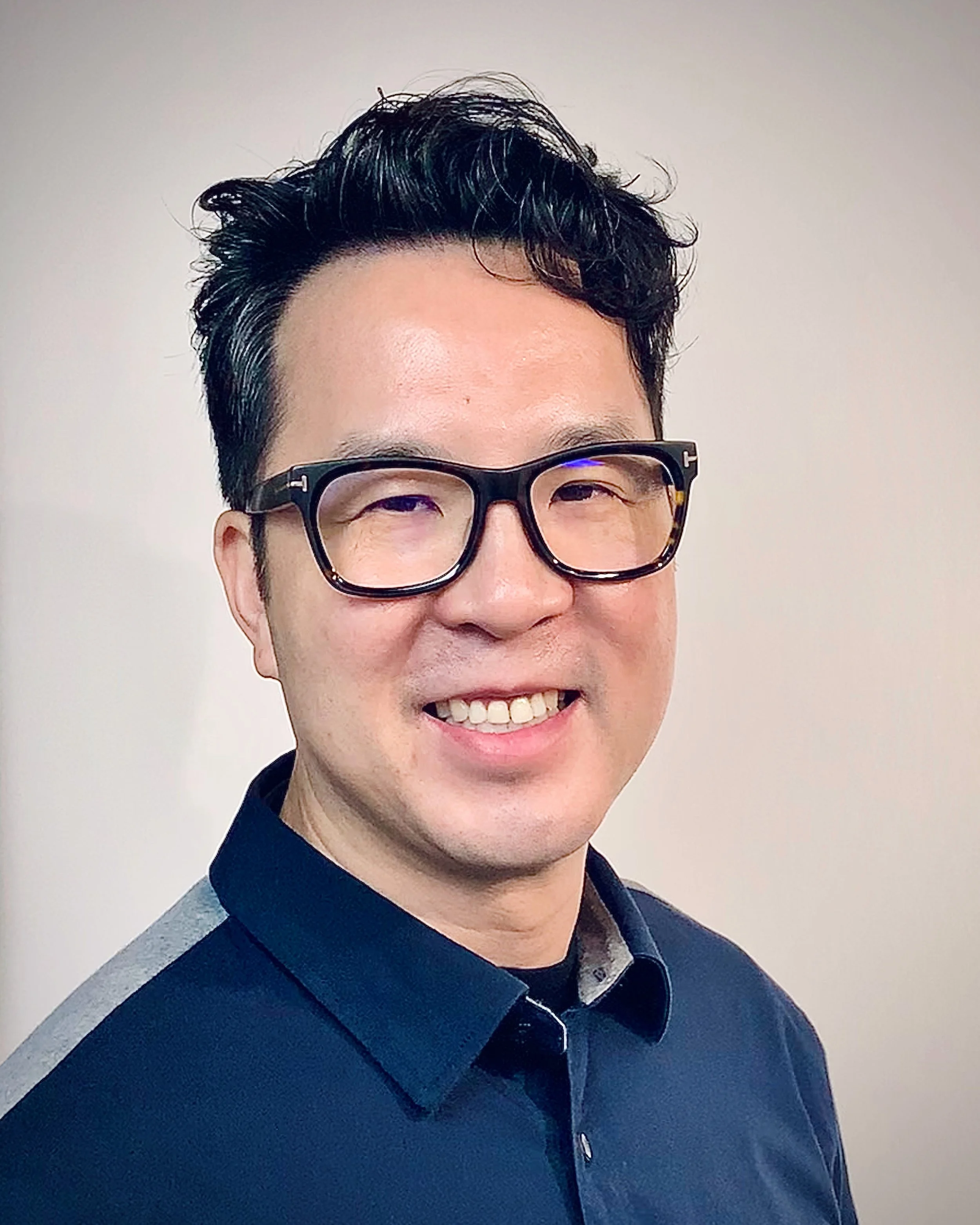 A smiling man wearing glasses and a collared shirt with a dark background.