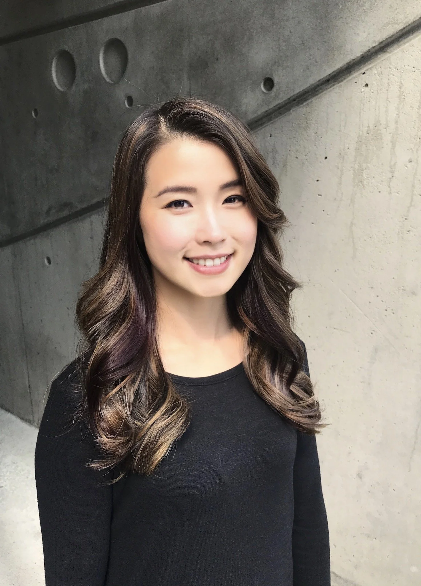 Smiling woman with long hair standing in front of a concrete wall.