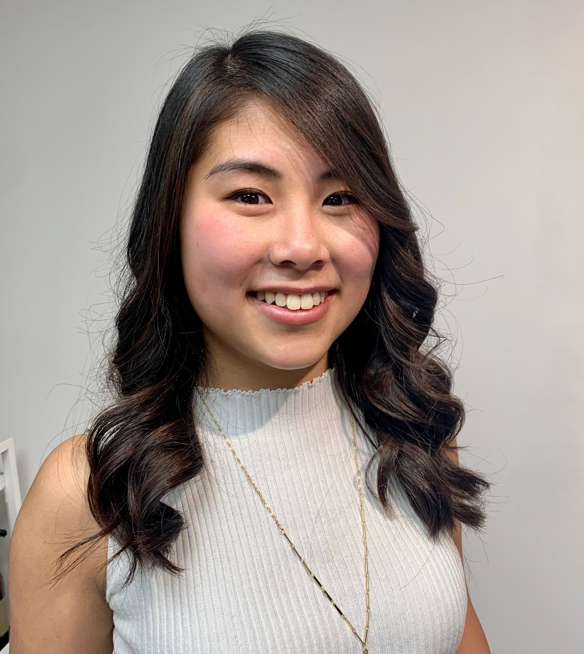A young woman smiling with wavy dark hair, wearing a sleeveless light-colored top and a gold necklace, standing against a plain background.