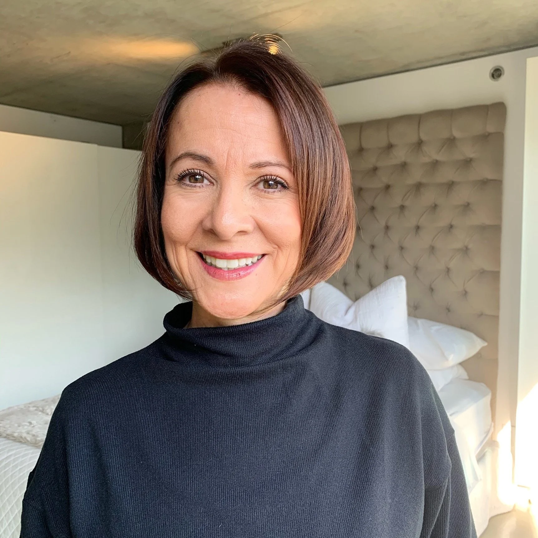 A smiling woman with shoulder-length brown hair, wearing a black turtleneck, standing in a bedroom with a tufted beige headboard and white pillows in the background.