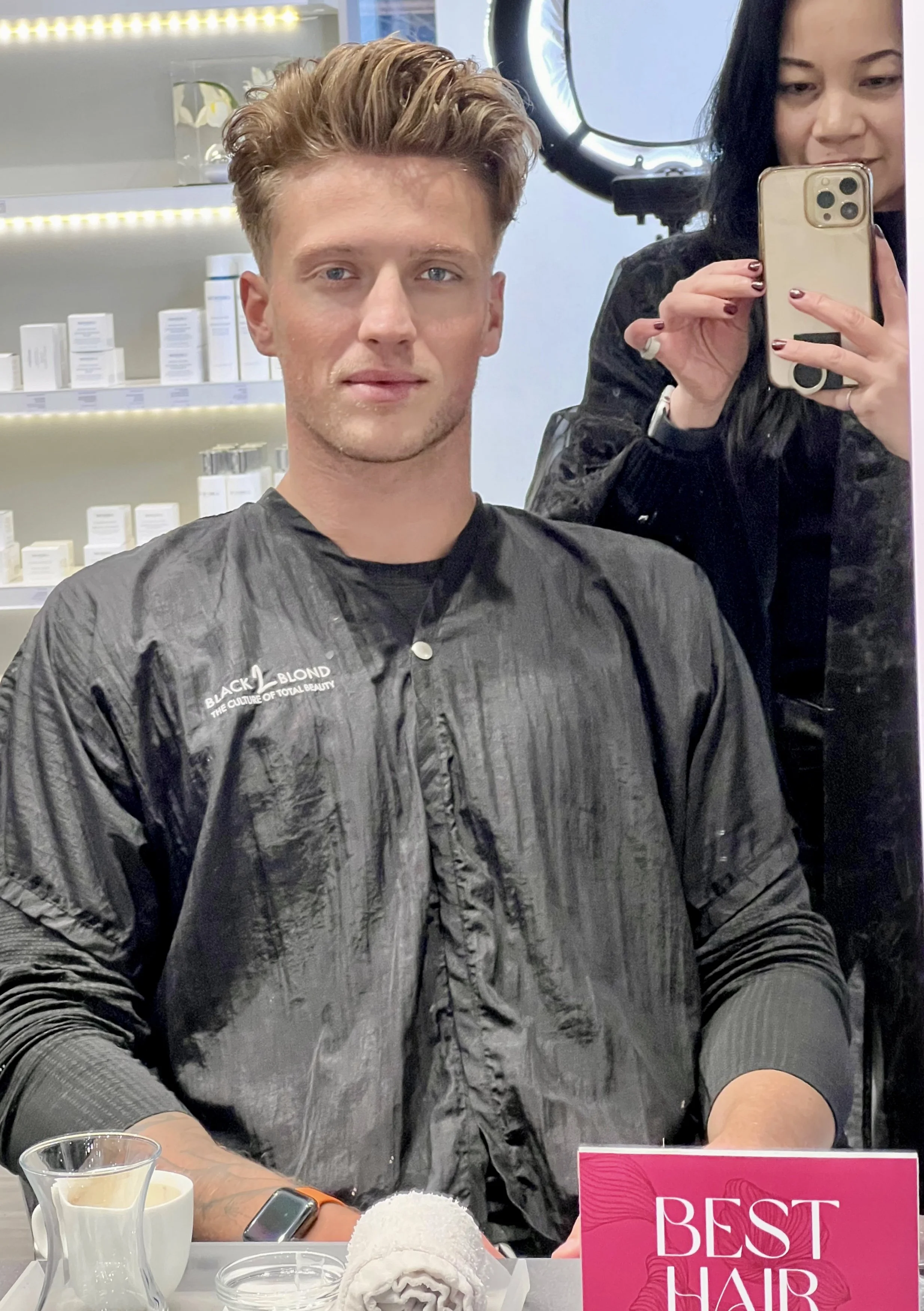 A man with styled hair sitting in a salon chair, wearing a black salon cape. A woman stands behind him holding a smartphone to take a photo. A pink sign that reads 'BEST HAIR' is visible on the table along with a cup of coffee, a rolled towel, and ha