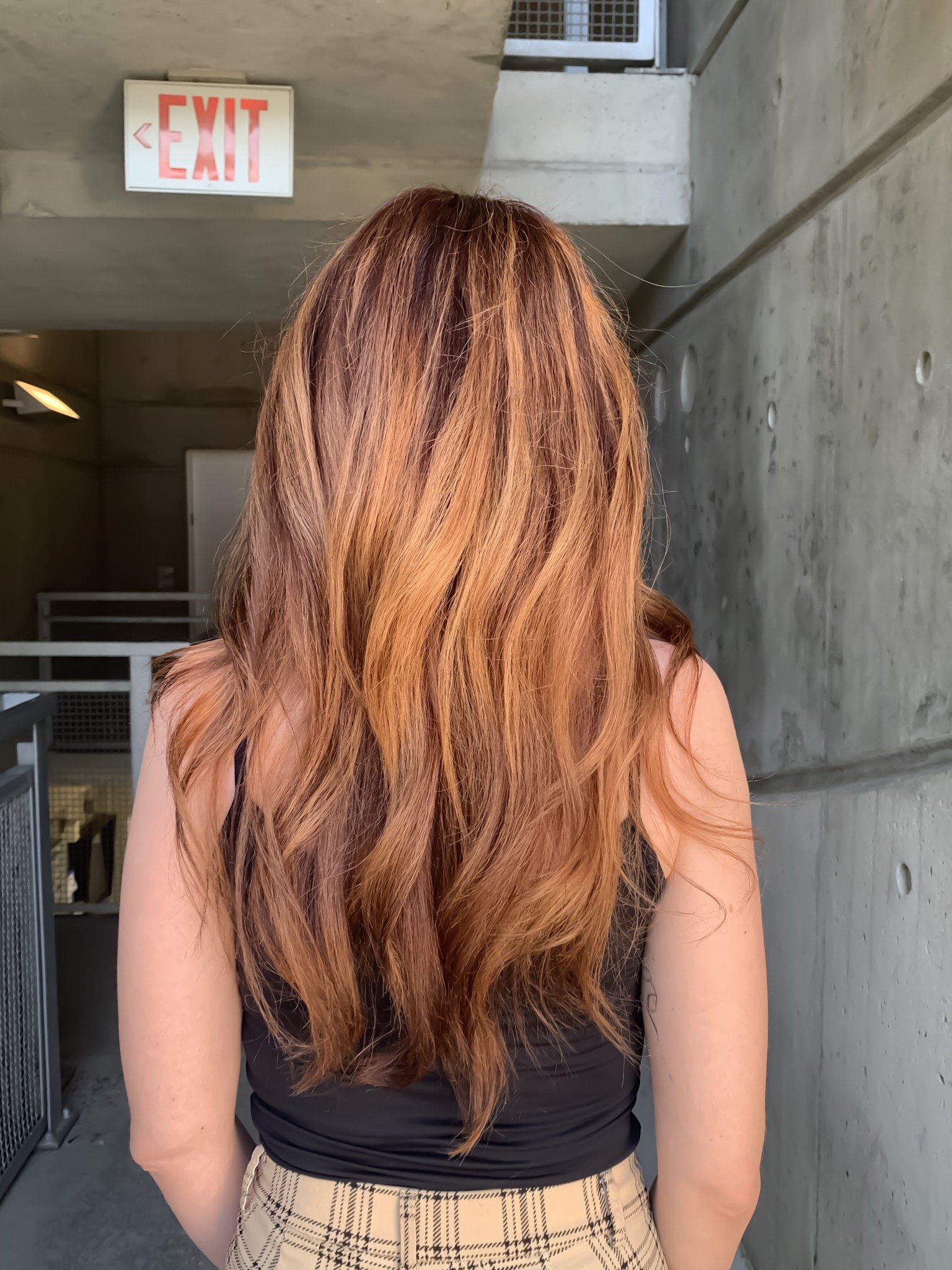 Person with long wavy auburn hair standing in front of a concrete wall, under an exit sign. Wearing a black top and plaid skirt.