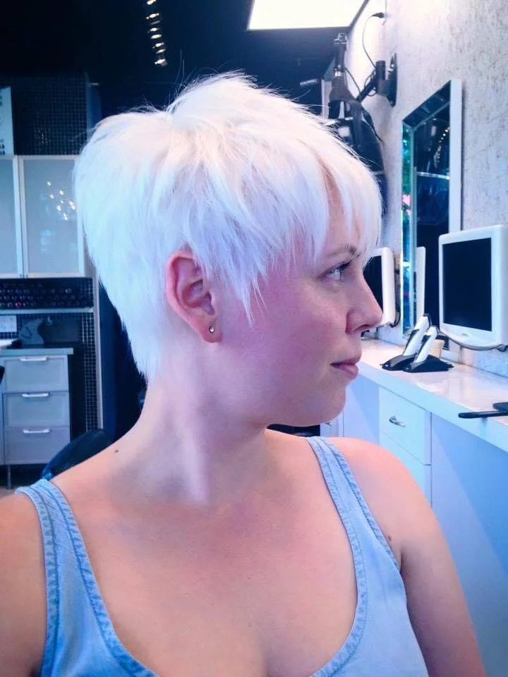 Short, platinum blonde hairstyle on a woman with earrings, sitting in a modern office or studio with computer monitors and equipment.