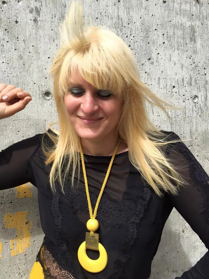 A woman with blonde hair and fair skin smiling with closed eyes, standing in front of a gray concrete wall, wearing a black top and a large yellow necklace.