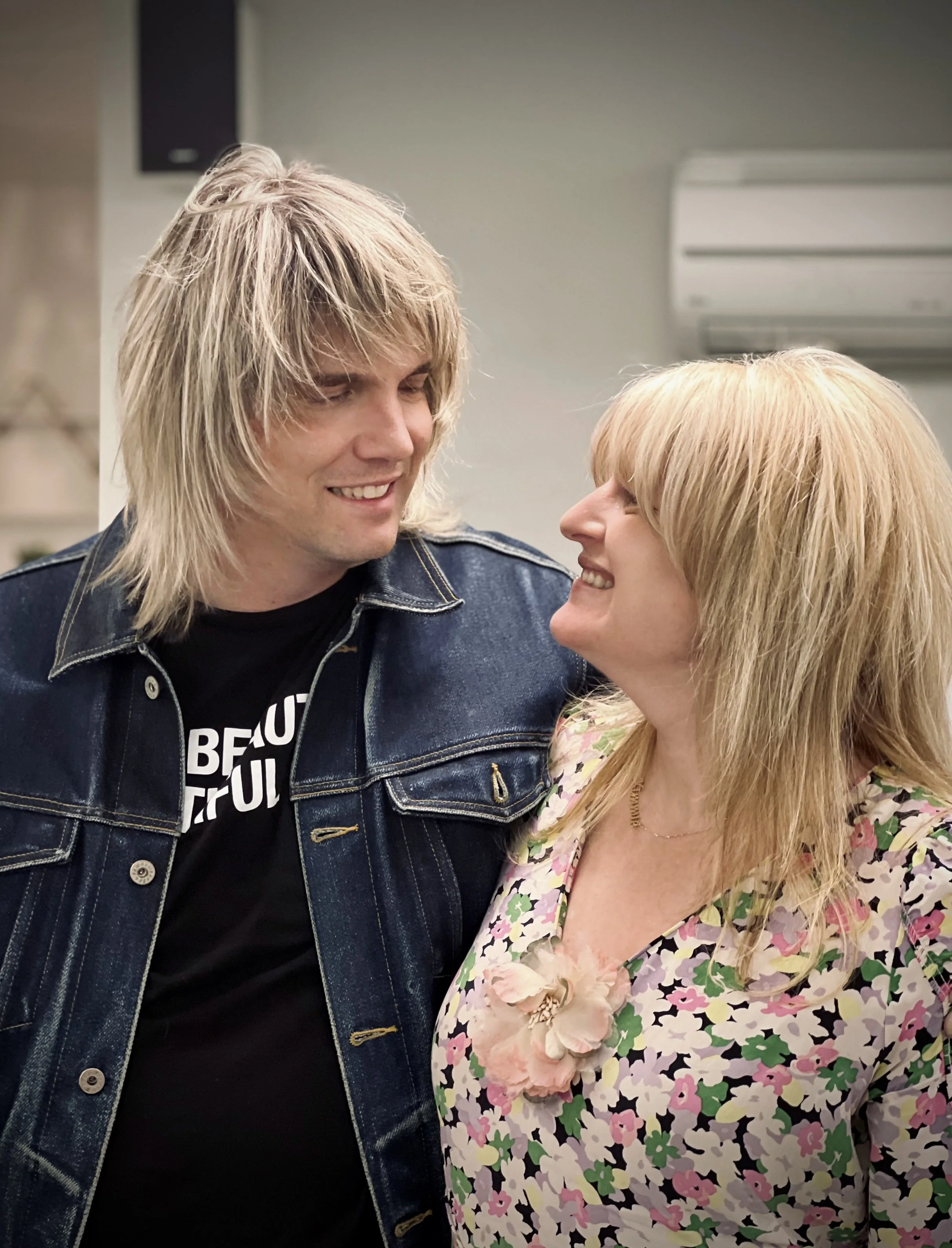 A couple with blonde hair smiling at each other indoors, with a man wearing a denim jacket and a woman wearing a floral dress with a flower accessory.