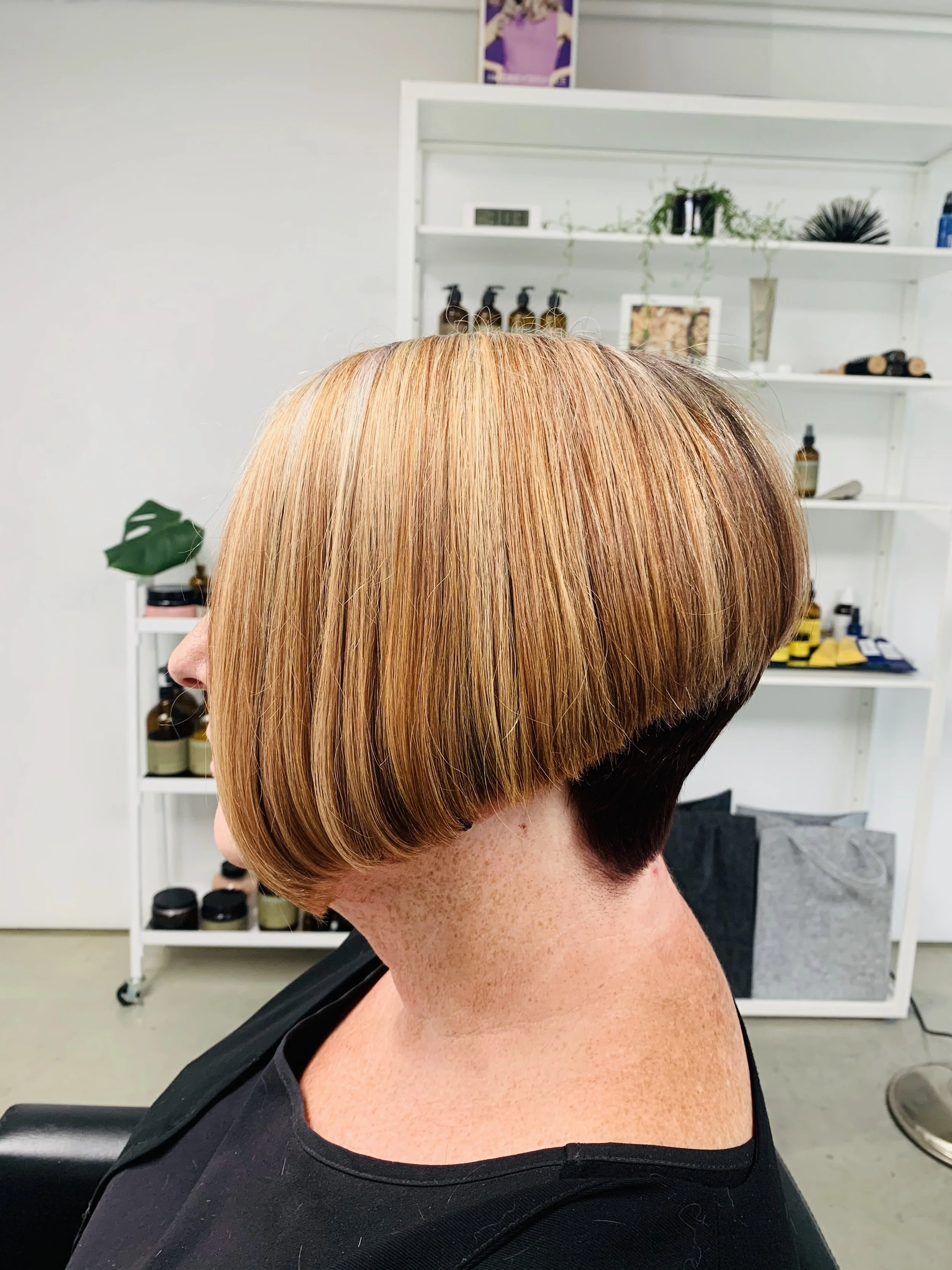 Side view of a woman with a short bob haircut with blonde highlights, sitting in a salon chair. Shelving in the background with various hair products.