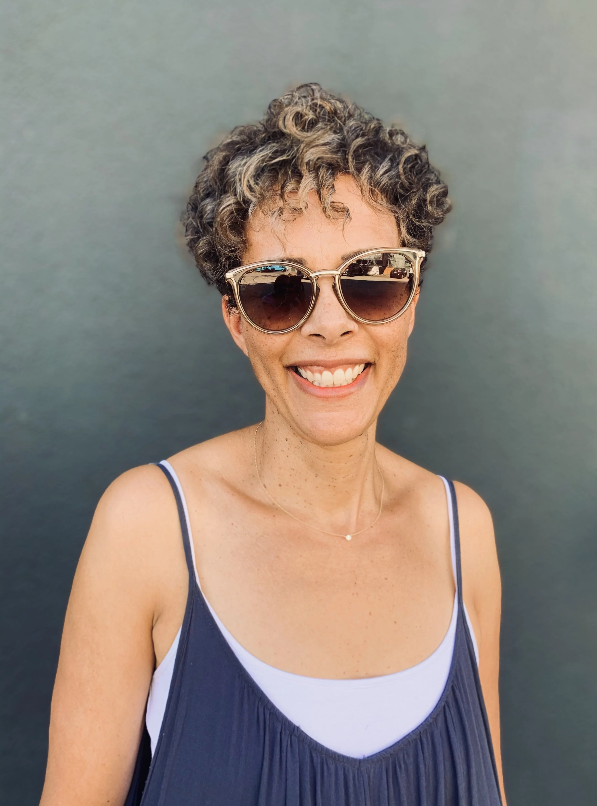Person with curly hair wearing sunglasses and a tank top, smiling against a plain background.