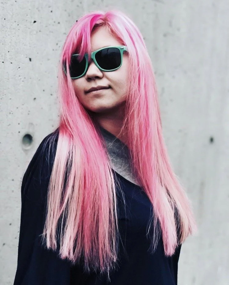 A woman with long pink hair wearing teal sunglasses and a dark jacket standing against a concrete wall.