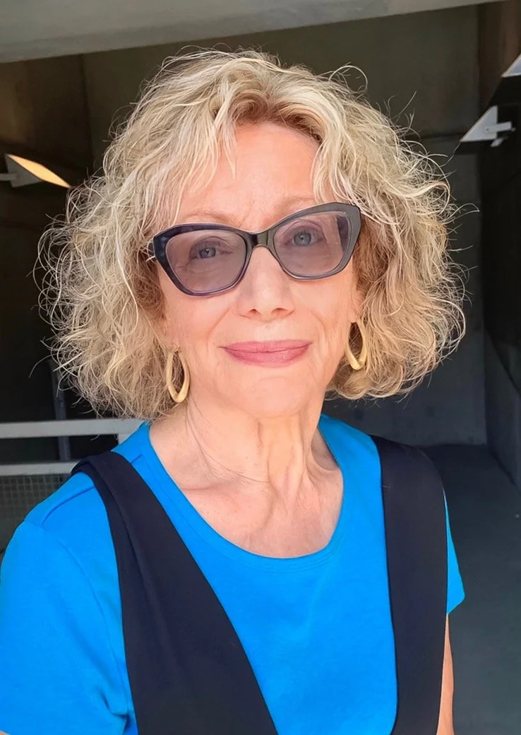 Portrait of a woman with curly blond hair, wearing sunglasses and a blue shirt with a black vest, smiling slightly, against a neutral background.