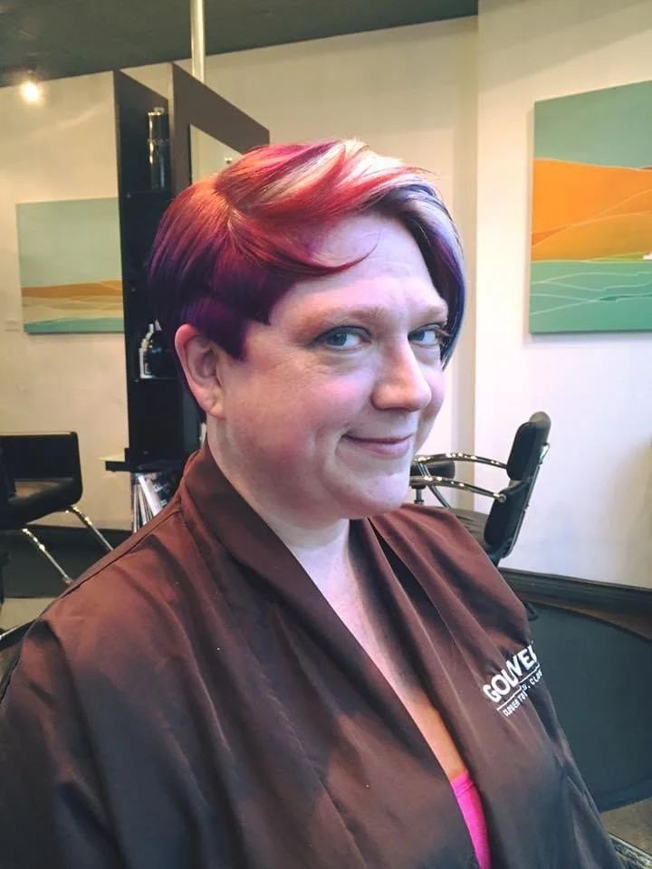 A woman with short, vibrant purple and red hair, wearing a brown salon cape, smiles at the camera inside a hair salon with modern decor, wall art, and salon chairs in the background.