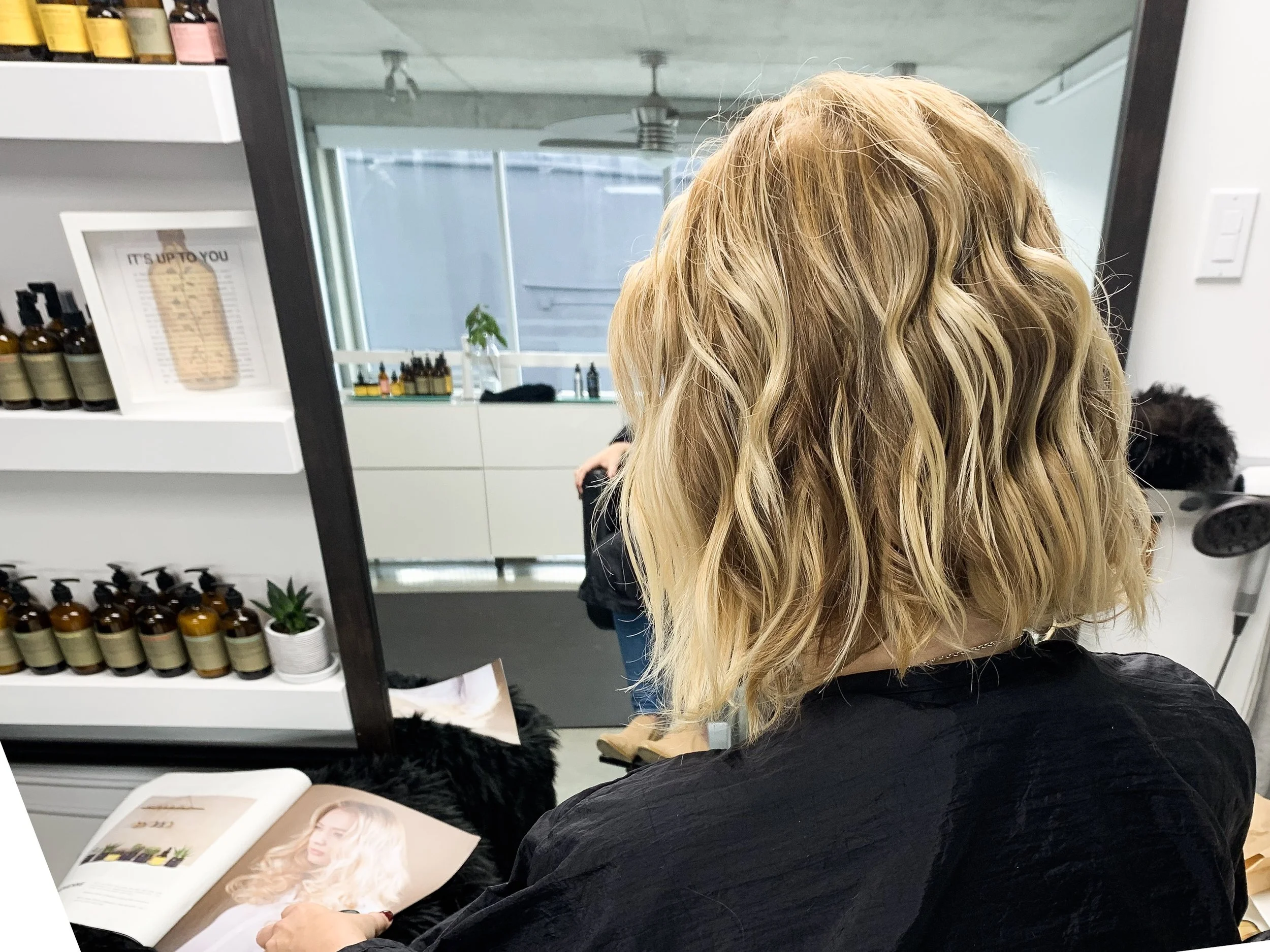 Person with wavy blonde hair in a salon in front of a mirror, holding a magazine, surrounded by hair products on shelves.