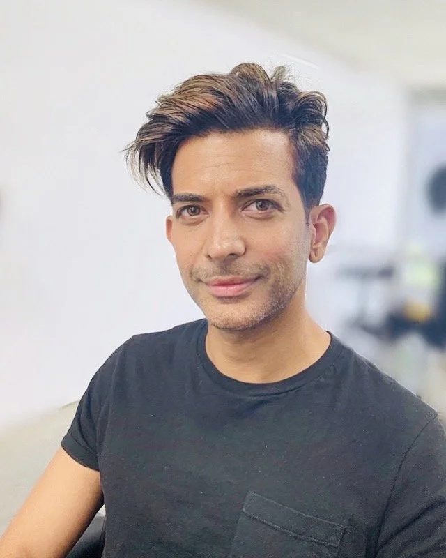A man with styled brown hair wearing a black t-shirt looking at the camera in a well-lit indoor setting.