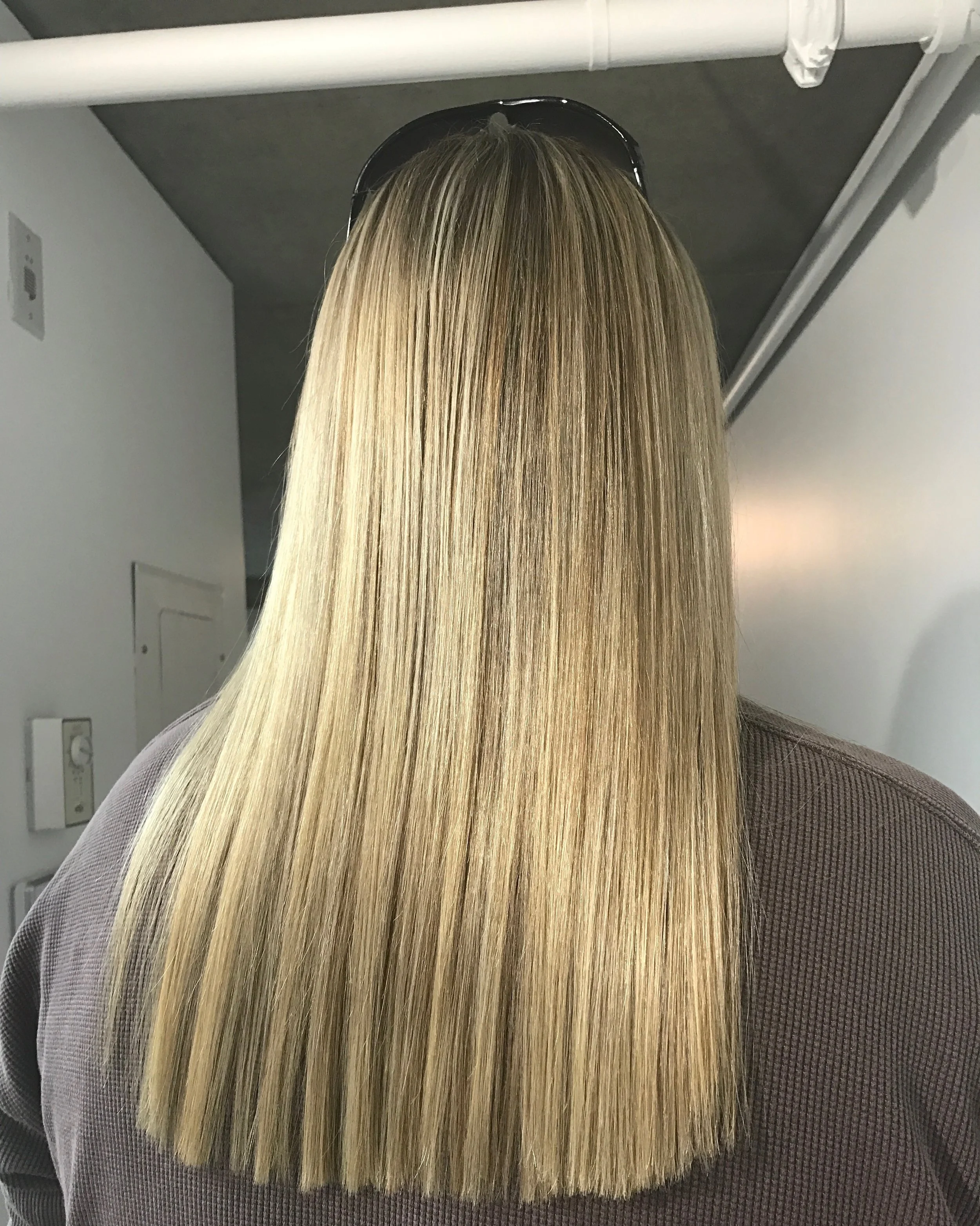 Back view of a woman with long, straight, blonde hair wearing sunglasses on her head and a gray top.