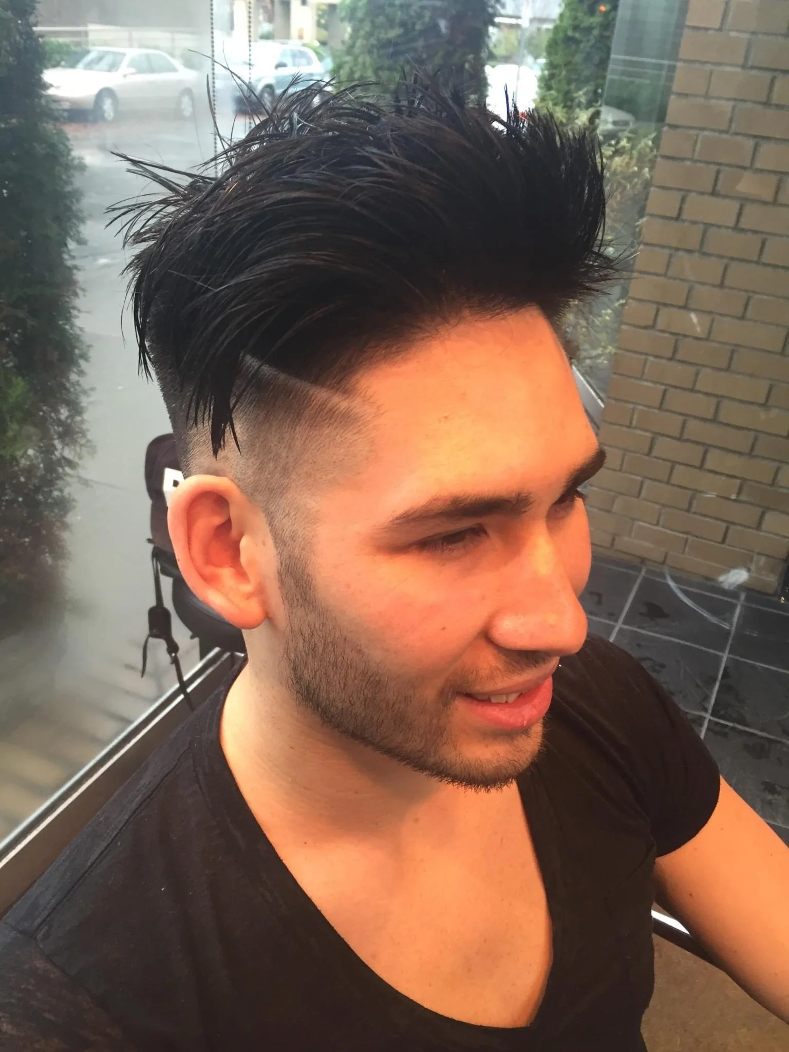 Young man with styled, spiked black hair and a fade haircut, sitting in a chair inside a barbershop.