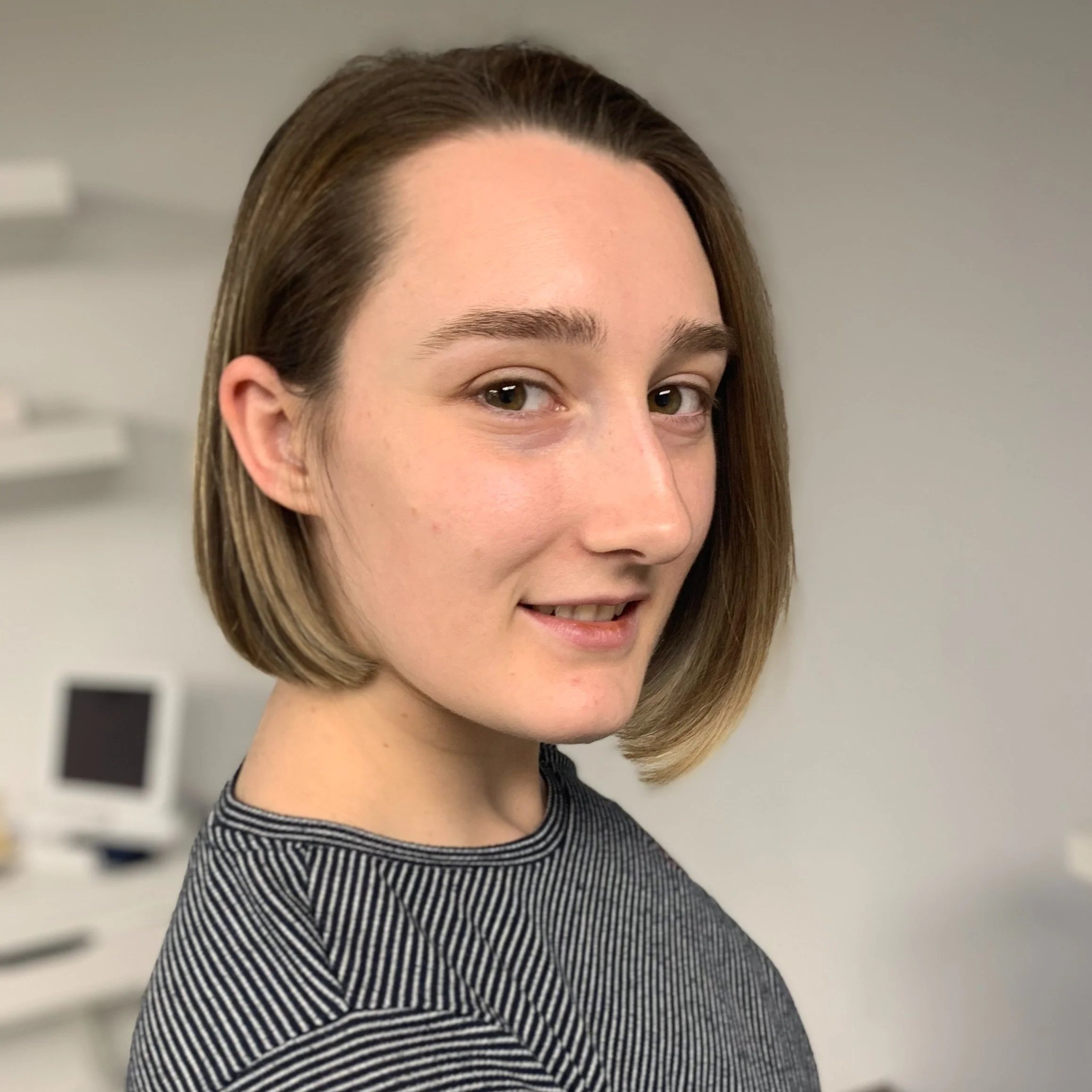 Close-up of a young woman with shoulder-length brown hair, smiling slightly, wearing a black-and-white striped top, in a room with medical equipment.