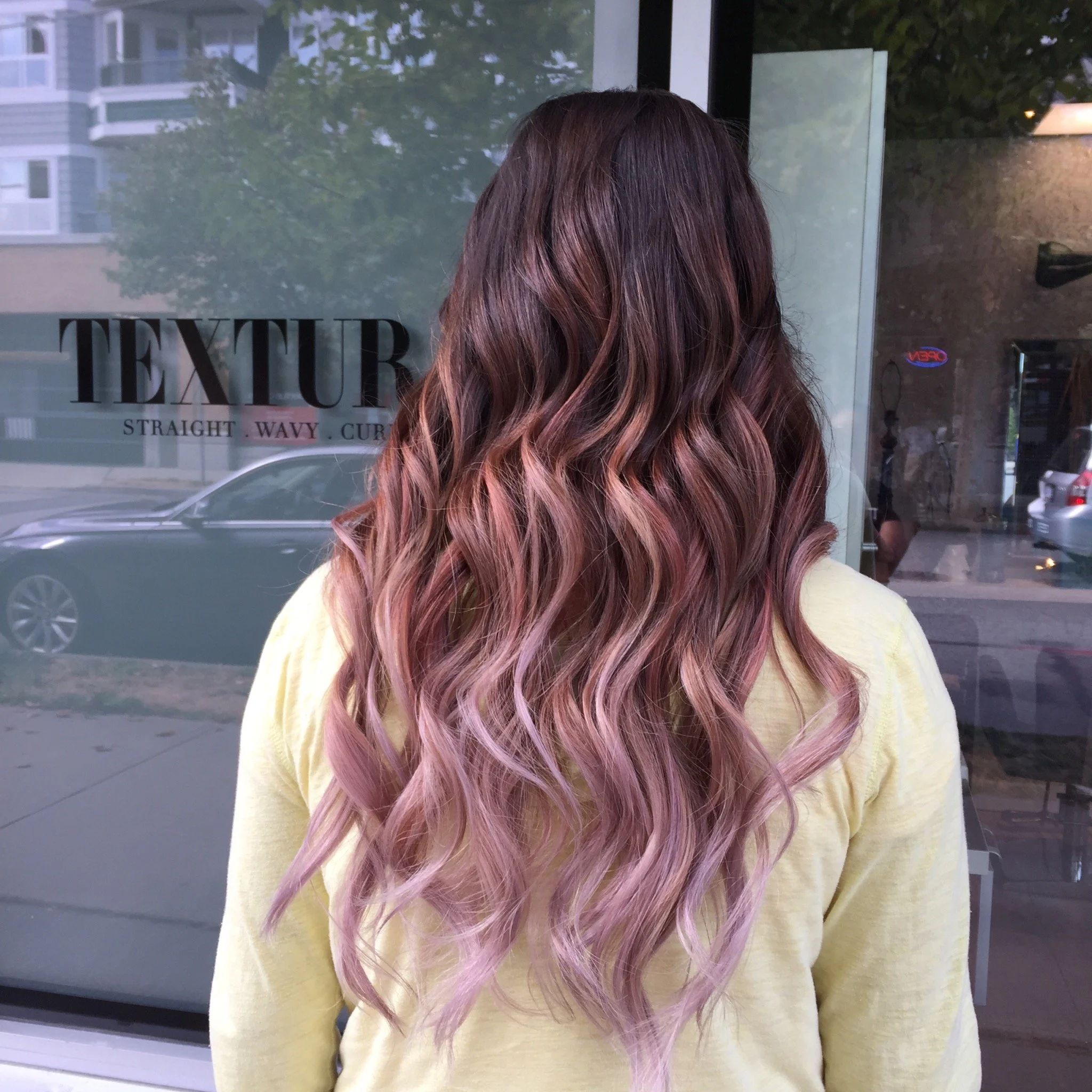 Woman with long, wavy balayage hair standing in front of hair salon window, wearing a yellow sweater.