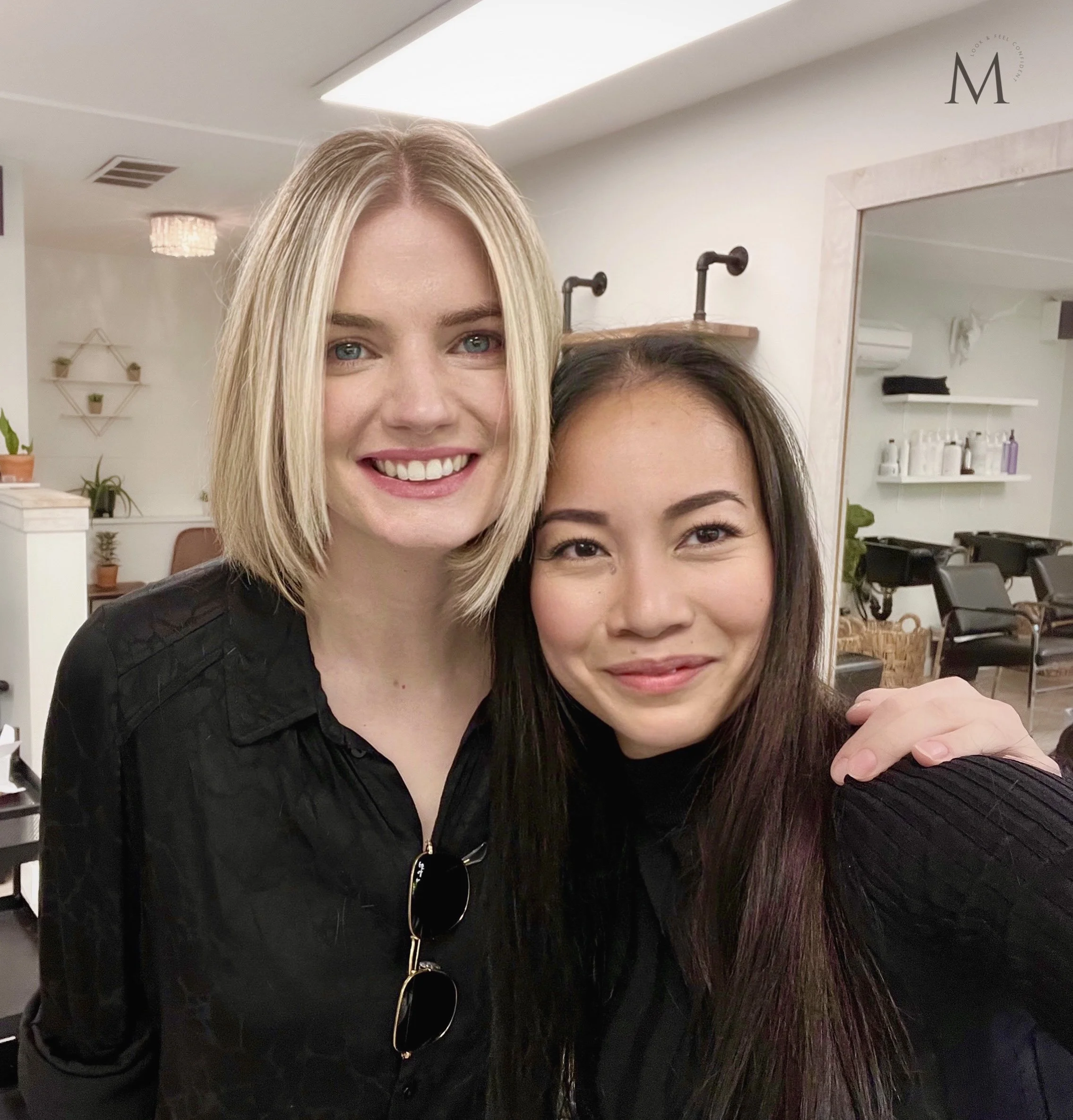 Two women smiling, standing in a hair salon, one with blonde hair and the other with dark hair.