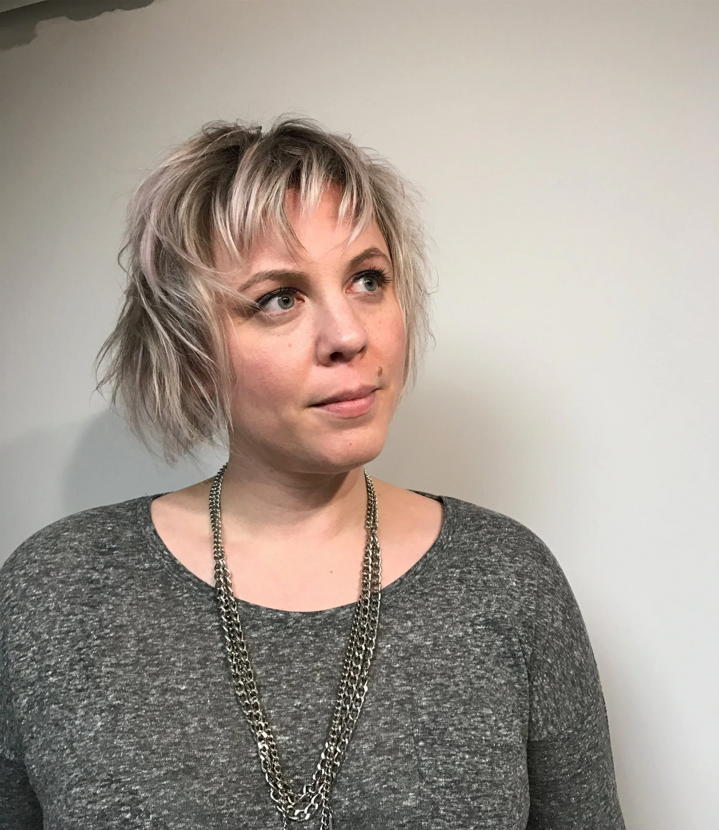 A woman with short, wavy blond hair and blue eyes, wearing a gray top and layered silver chains, stands against a plain off-white background.