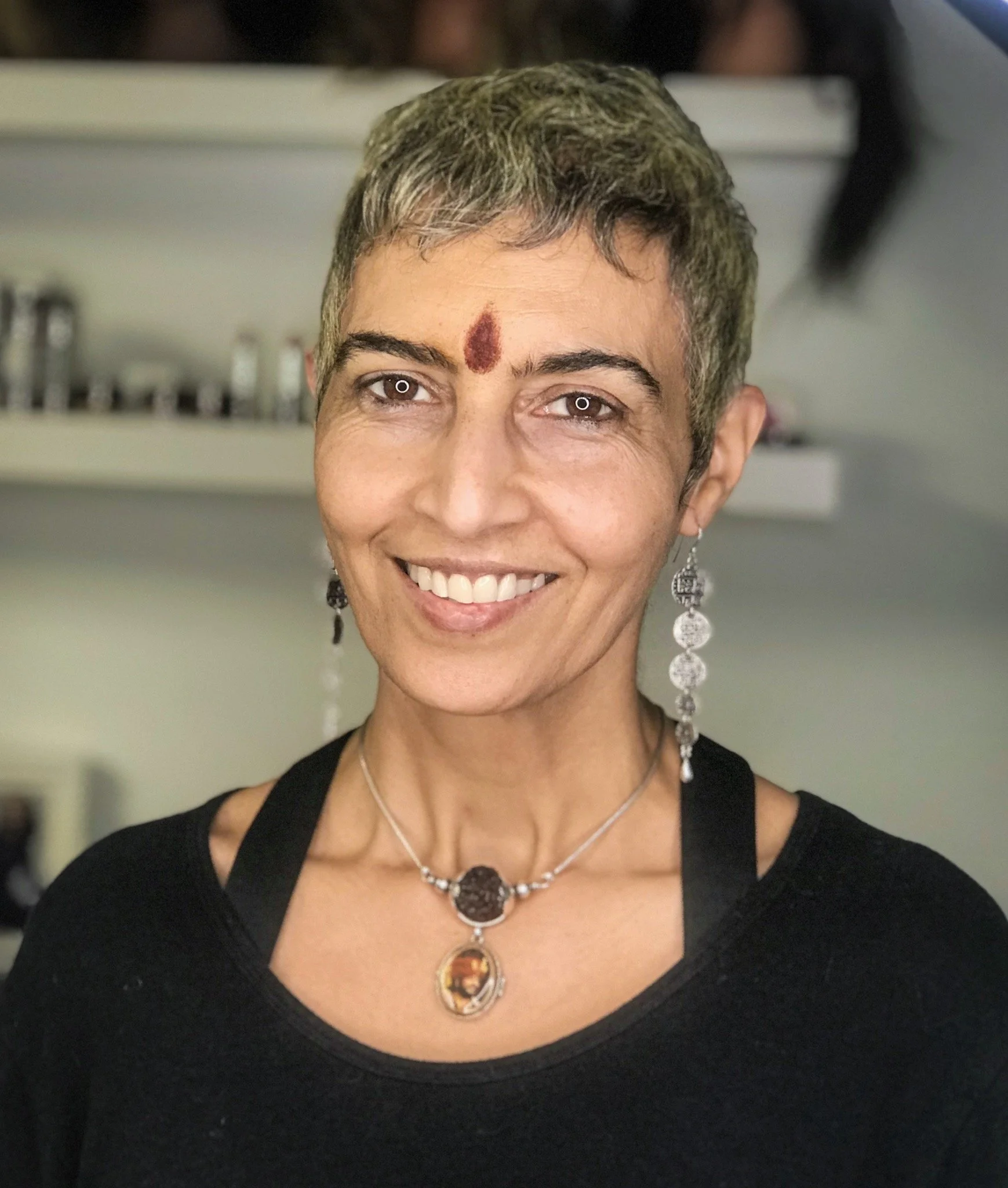 A smiling woman with short gray hair, wearing earrings and a necklace, with a red mark on her forehead.
