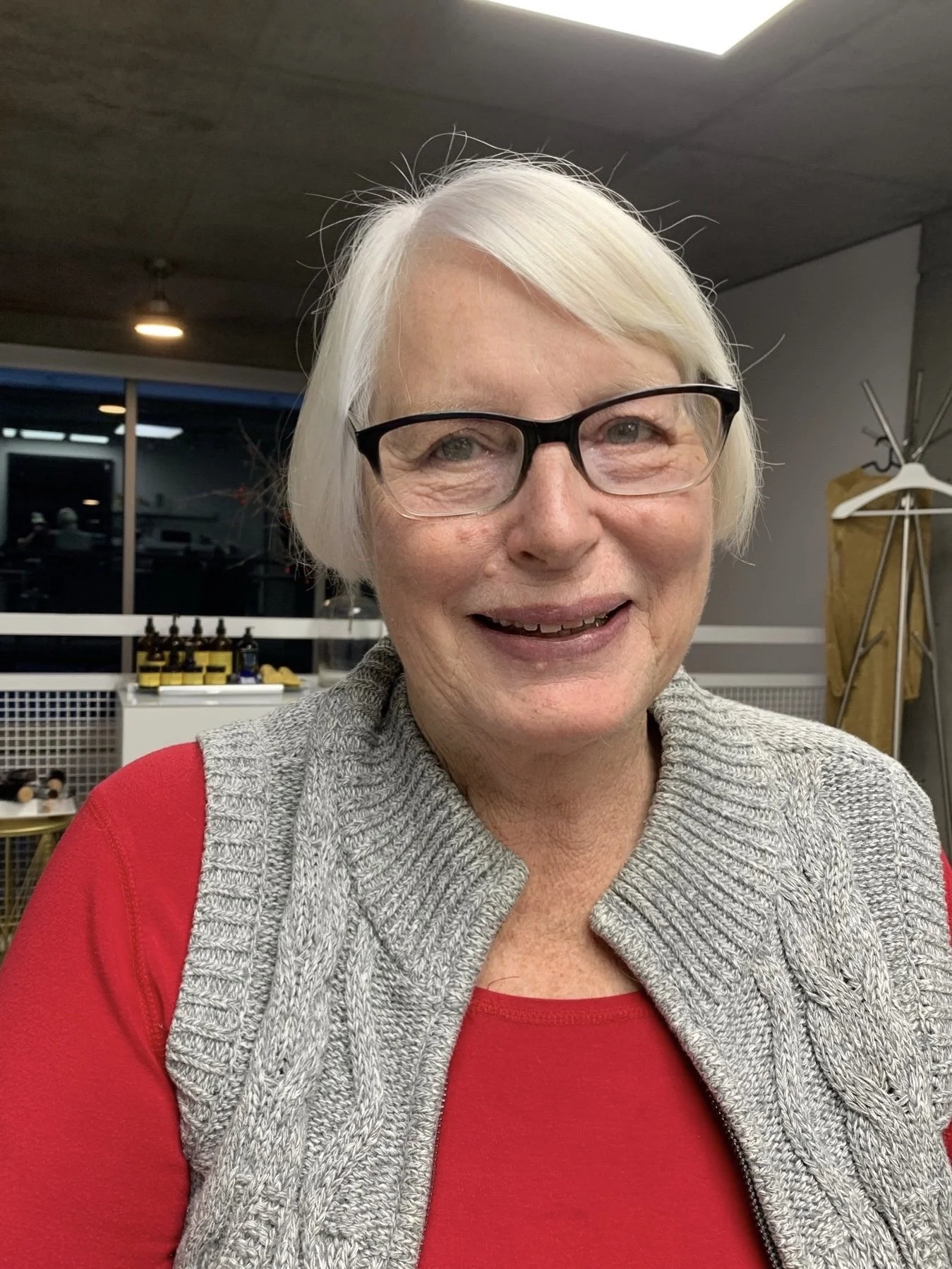 A smiling older woman with short white hair, wearing glasses, a red shirt, and a gray knitted vest, indoors with cancer care products in the background.