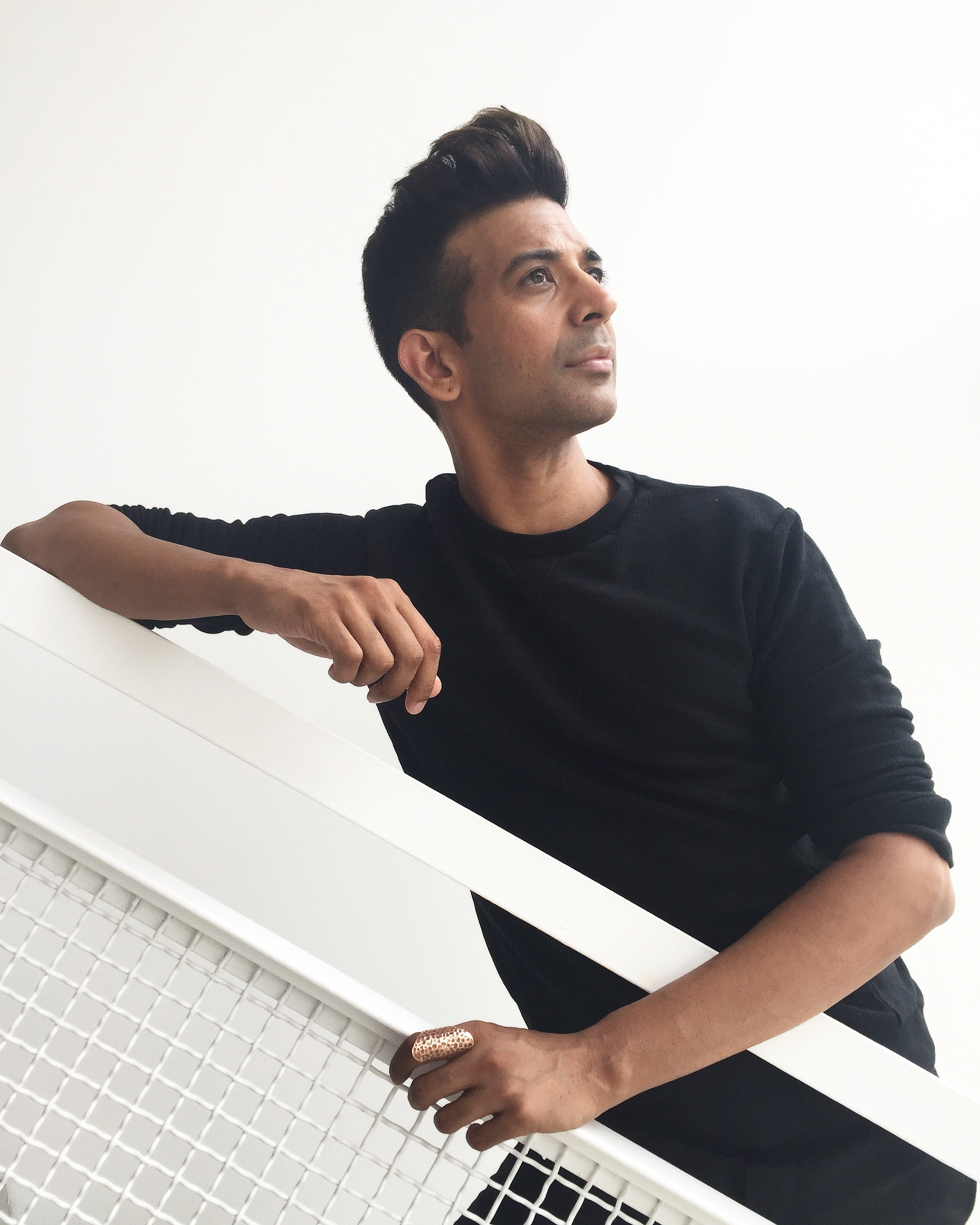 A man with dark hair styled upwards, wearing a black shirt, leaning on a white railing with a white background.
