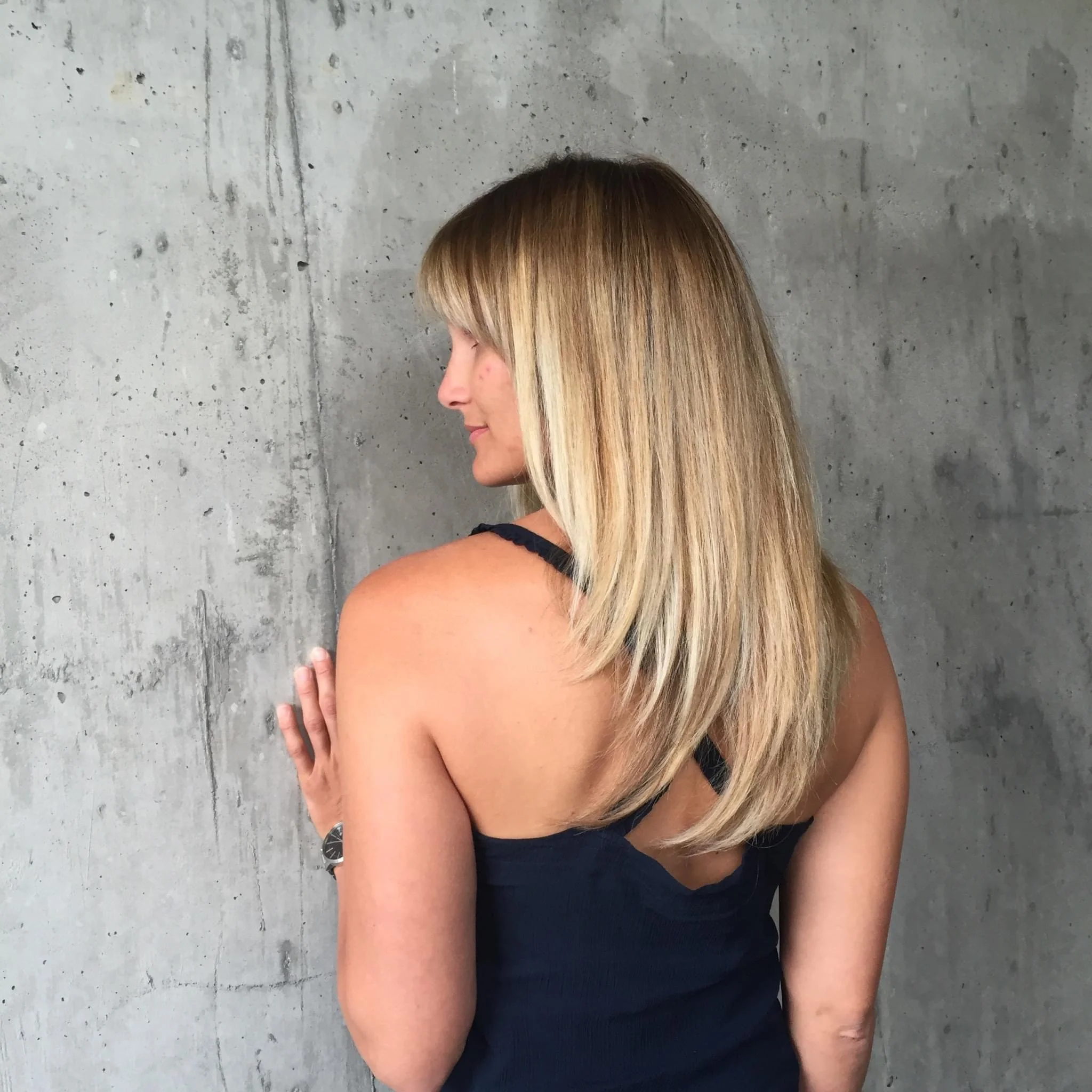 Side view of a woman with long blonde hair wearing a black dress, standing against a concrete wall.