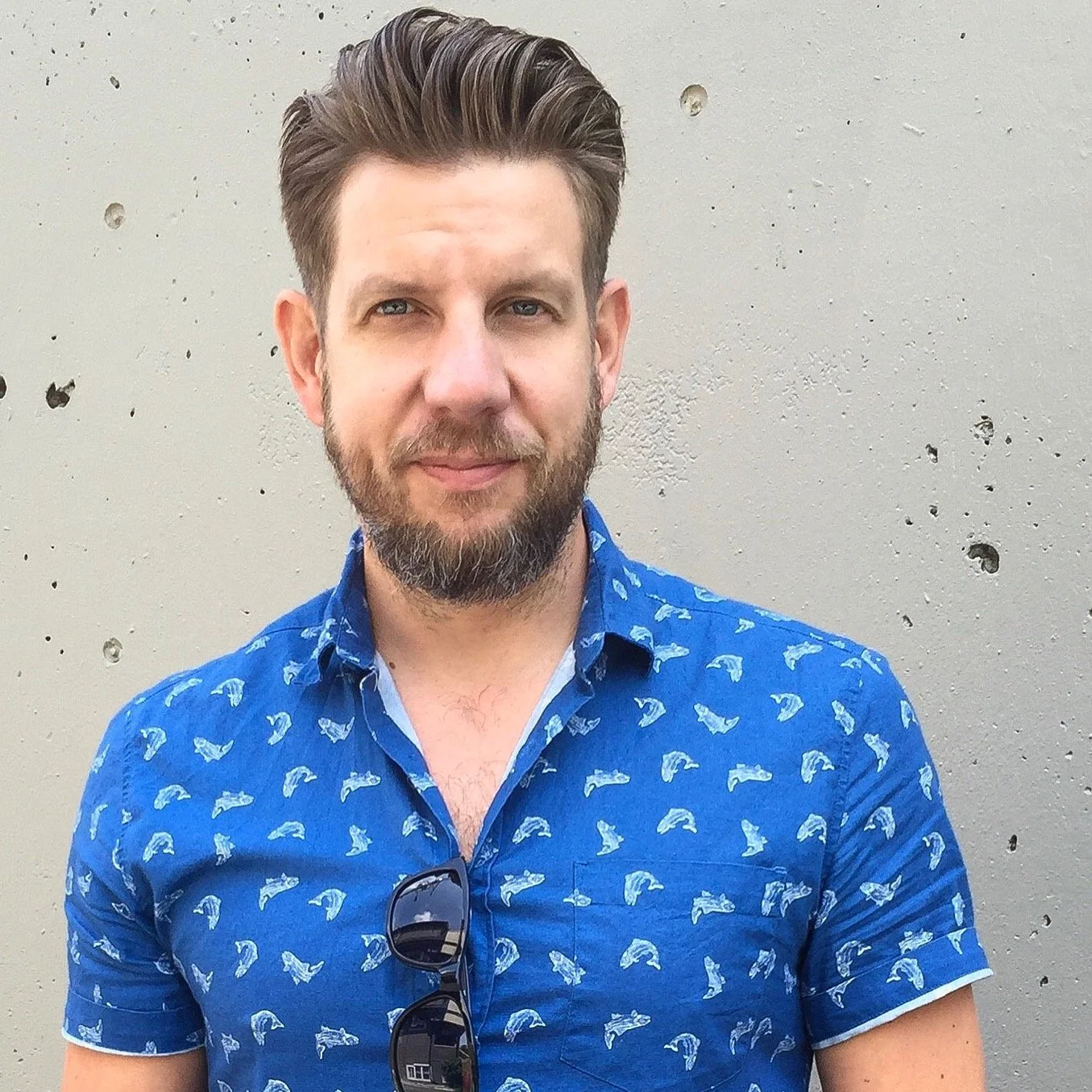 Man with brown hair and beard wearing a blue shirt with whale print, standing in front of a beige concrete wall.